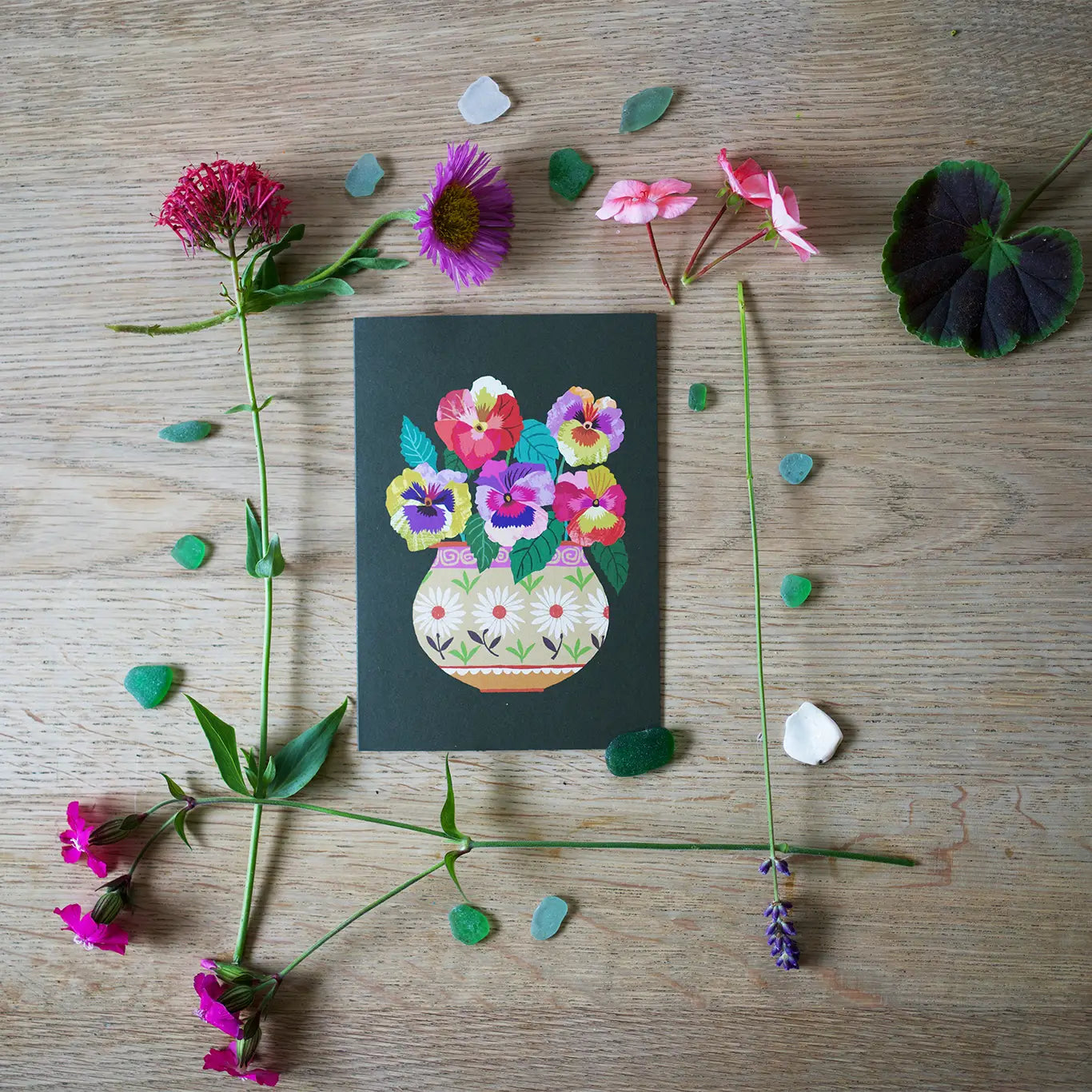 Decorative card with floral design surrounded by real flowers and leaves on a wooden surface.
