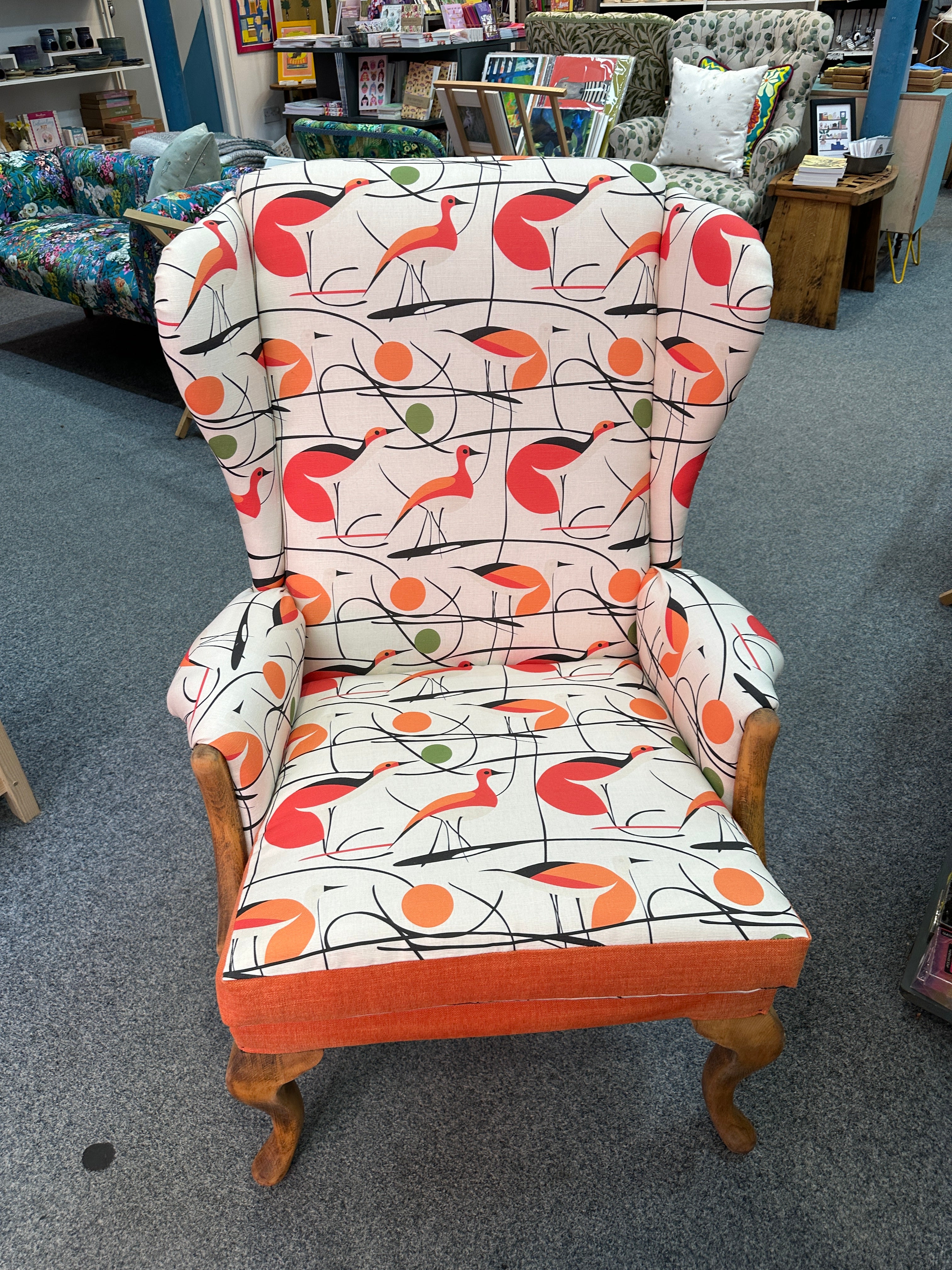 An armchair with a retro mid-century modern bird pattern on a white background, featuring orange, red, and green birds with black lines, and orange velvet trim on the arms and backrest.