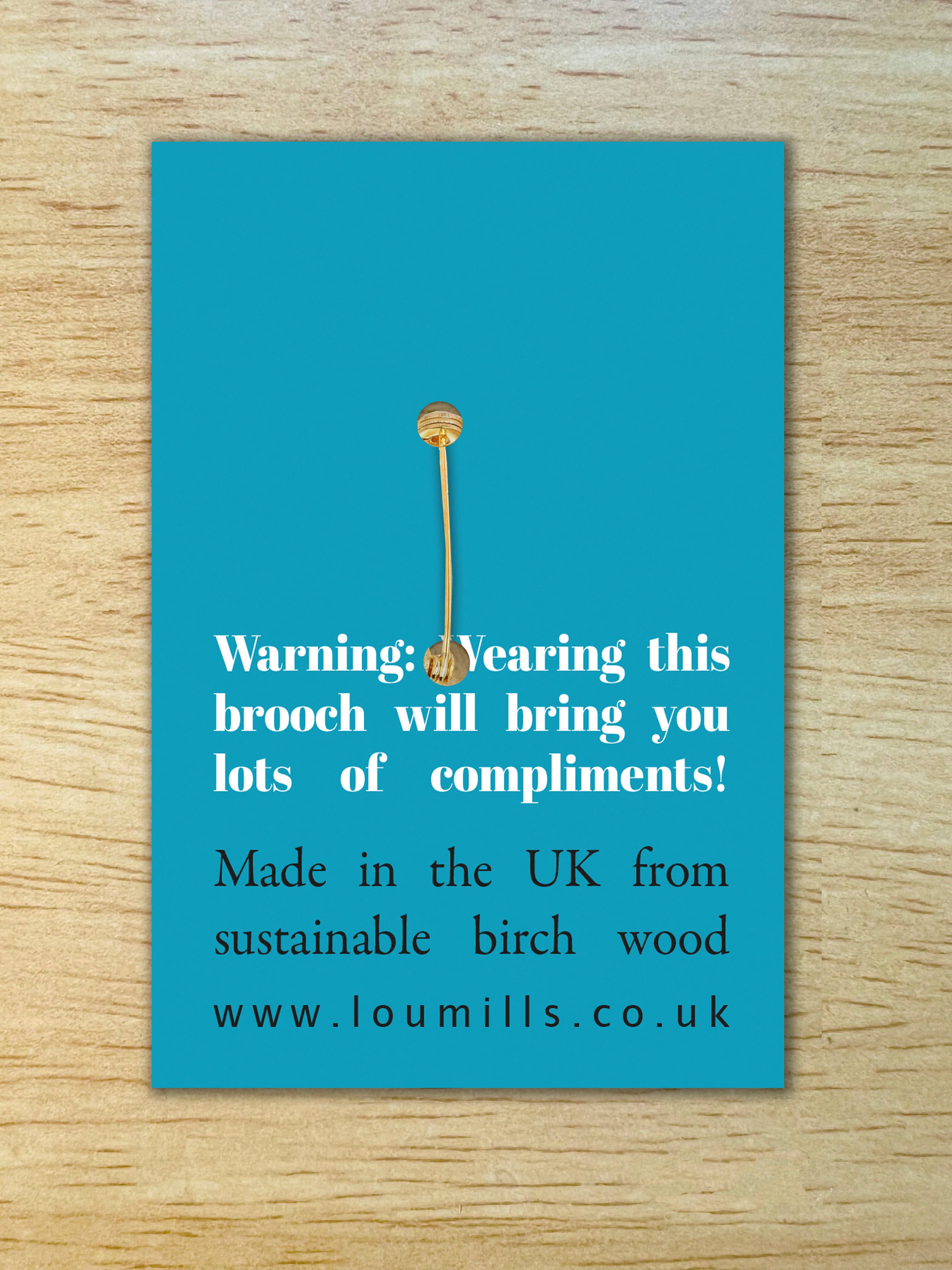 A single gold-toned nail stud brooch is centred on a bright turquoise rectangular card with white and dark text, placed on a light wood surface.