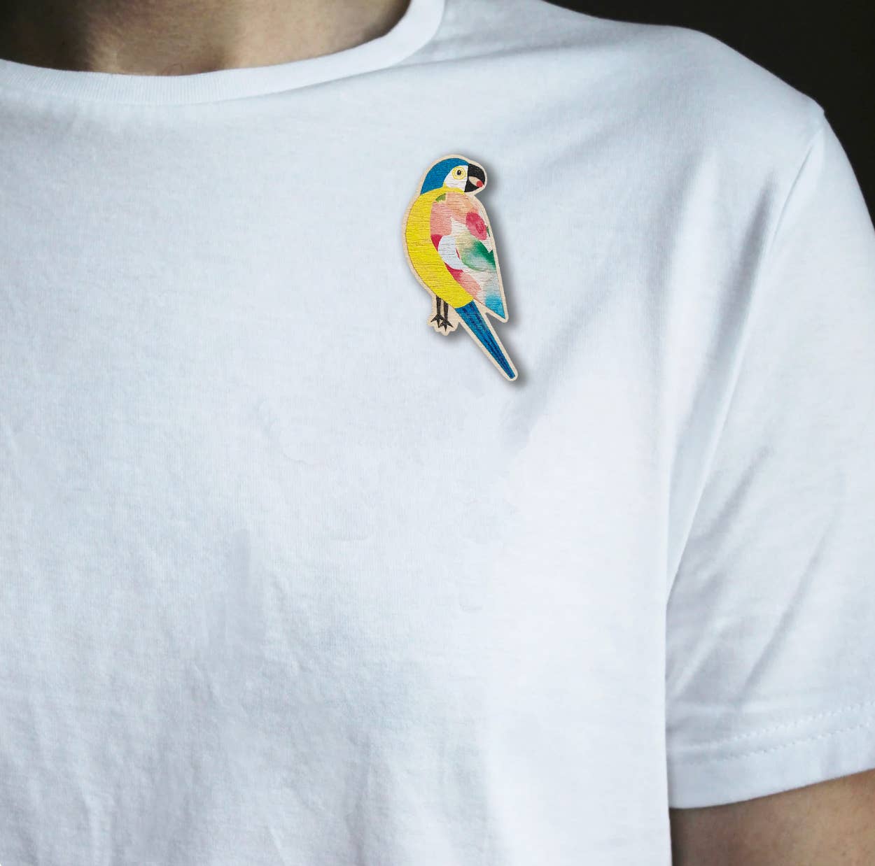 A close-up of a person wearing a white crew-neck t-shirt with a colorful parrot enamel pin on the left chest.