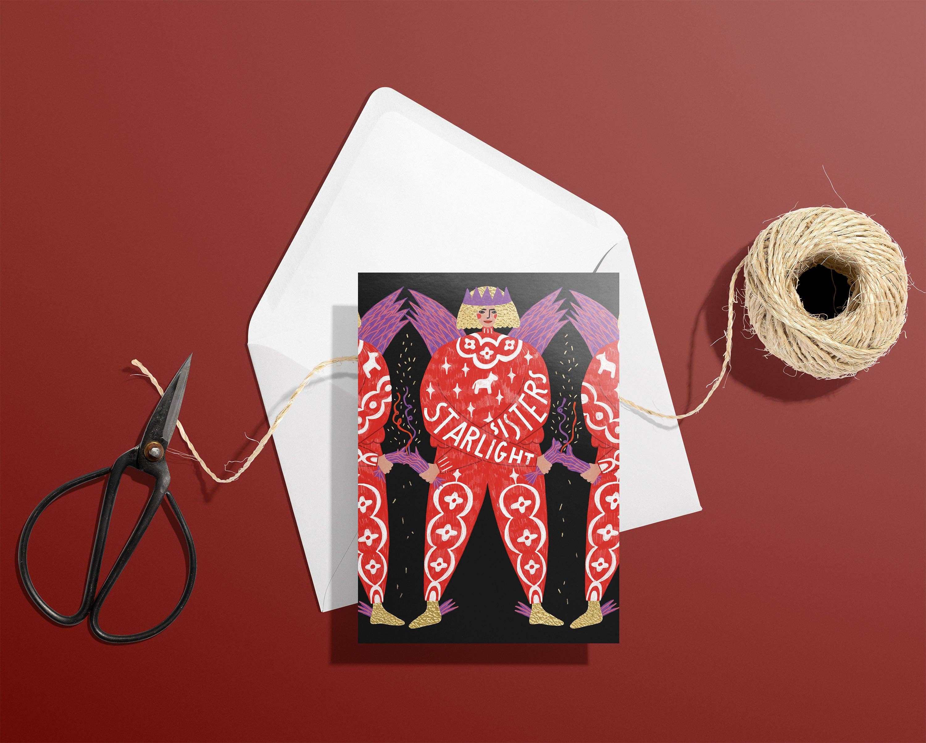 A square greeting card with a stylised red and purple figure and the word 'STARLIGHT SISTERS' is layered over a white envelope, arranged on a dark red background with black scissors and a spool of twine.