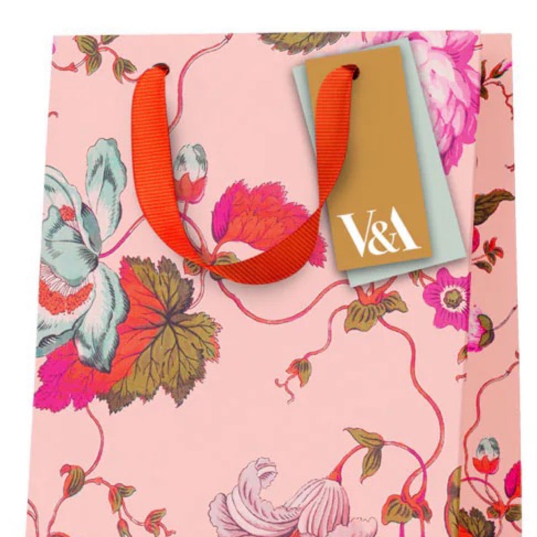 Colourful gift bag featuring pink, white and red flowers against a peach background and a red ribbon handle.