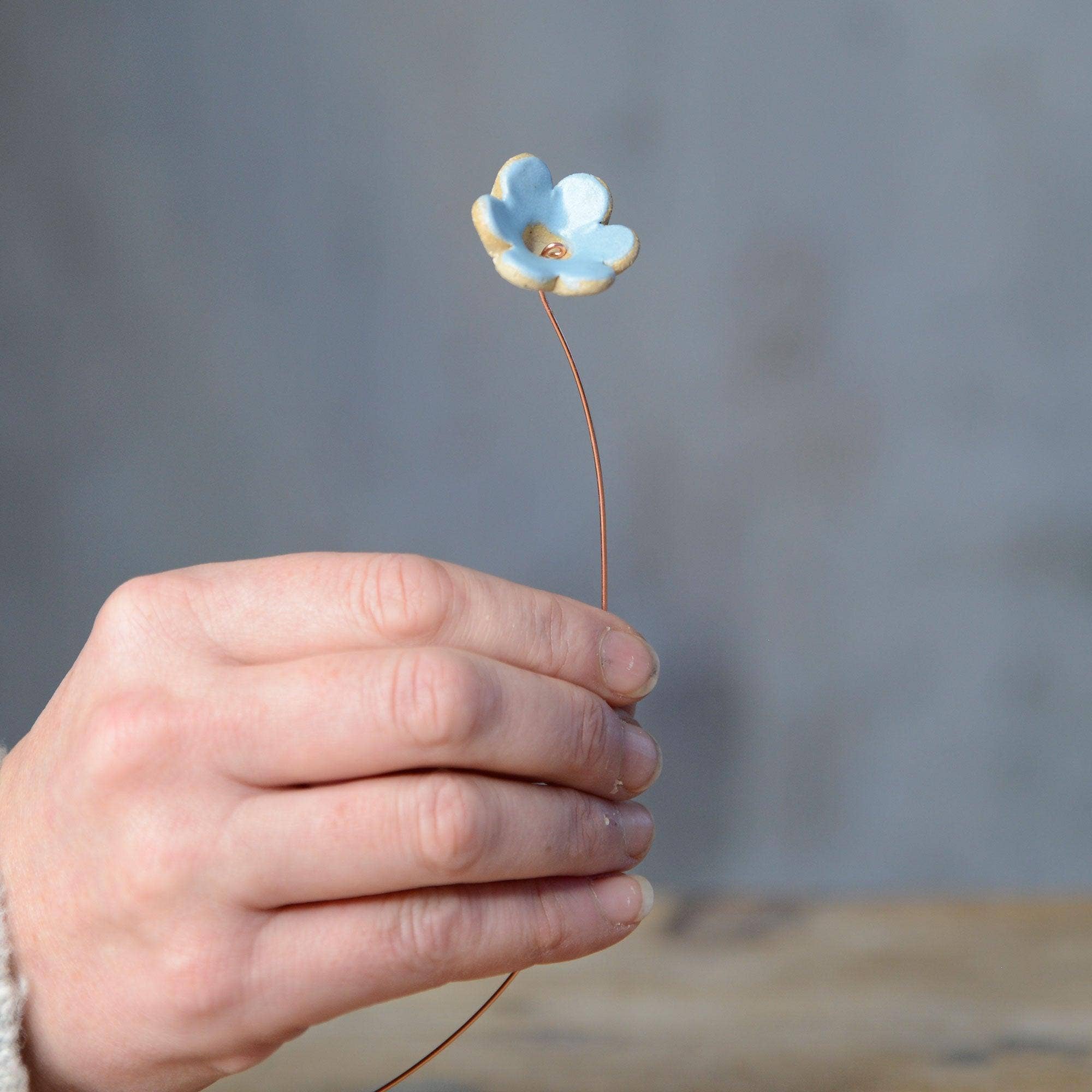 A hand holding a pastel blue ceramic flower bud stem on a copper wire