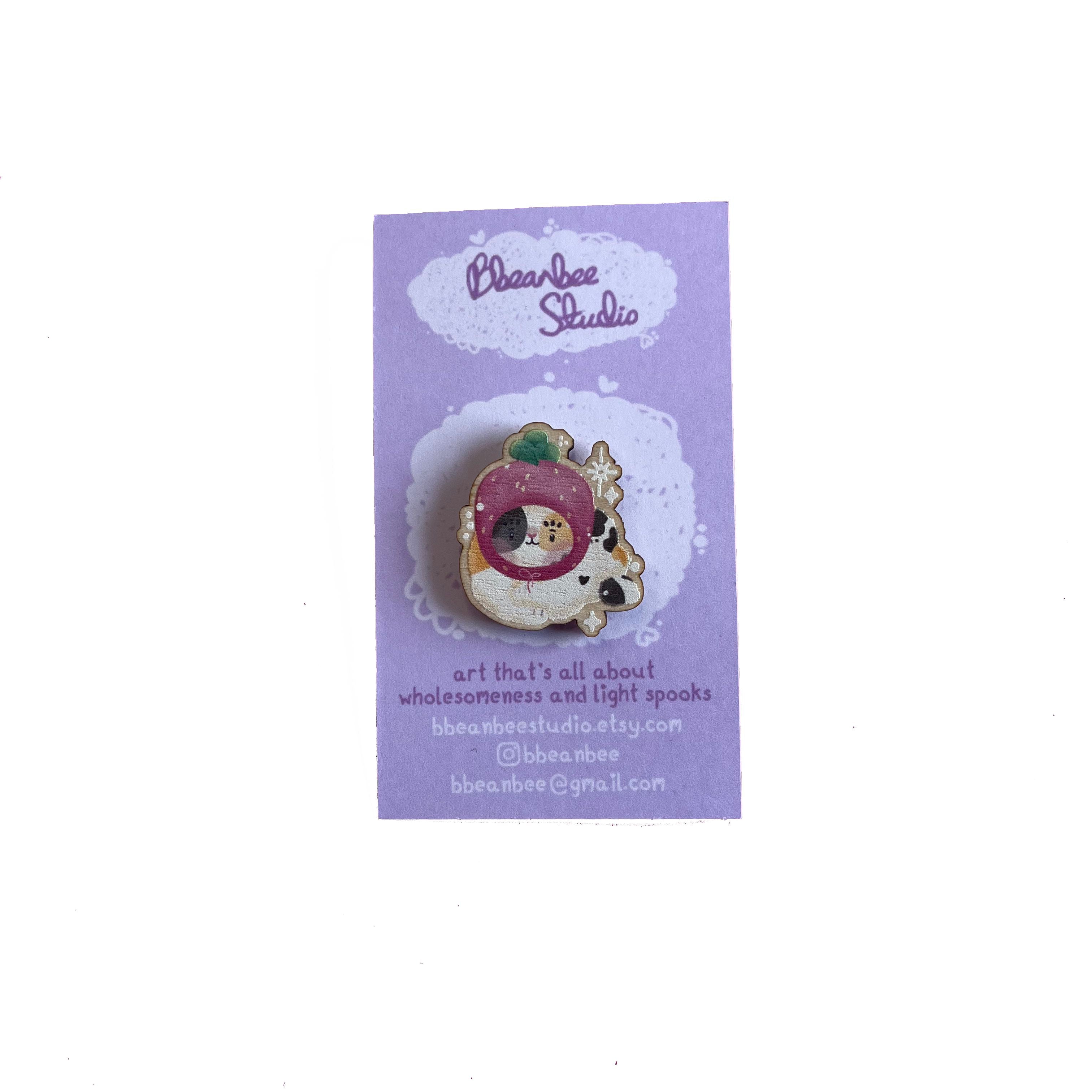 Wooden pin badge of a calico cat wearing a pawberry (heart-shaped strawberries with a paw shape inside) hat. This badge is printed onto 3mm thick responsibly sourced European birch plywood.
