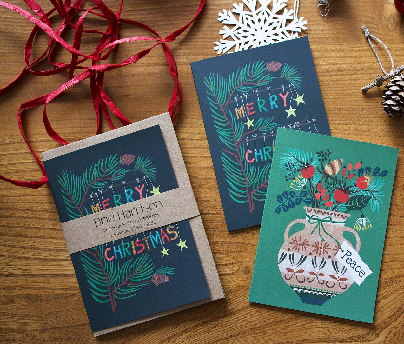 Three Christmas greeting cards arranged on a wooden surface with red ribbon, a white snowflake ornament, and a pinecone. One card is wrapped in a kraft paper band reading 'Merry Christmas' and the card with 'Brie Harrison' packaging.