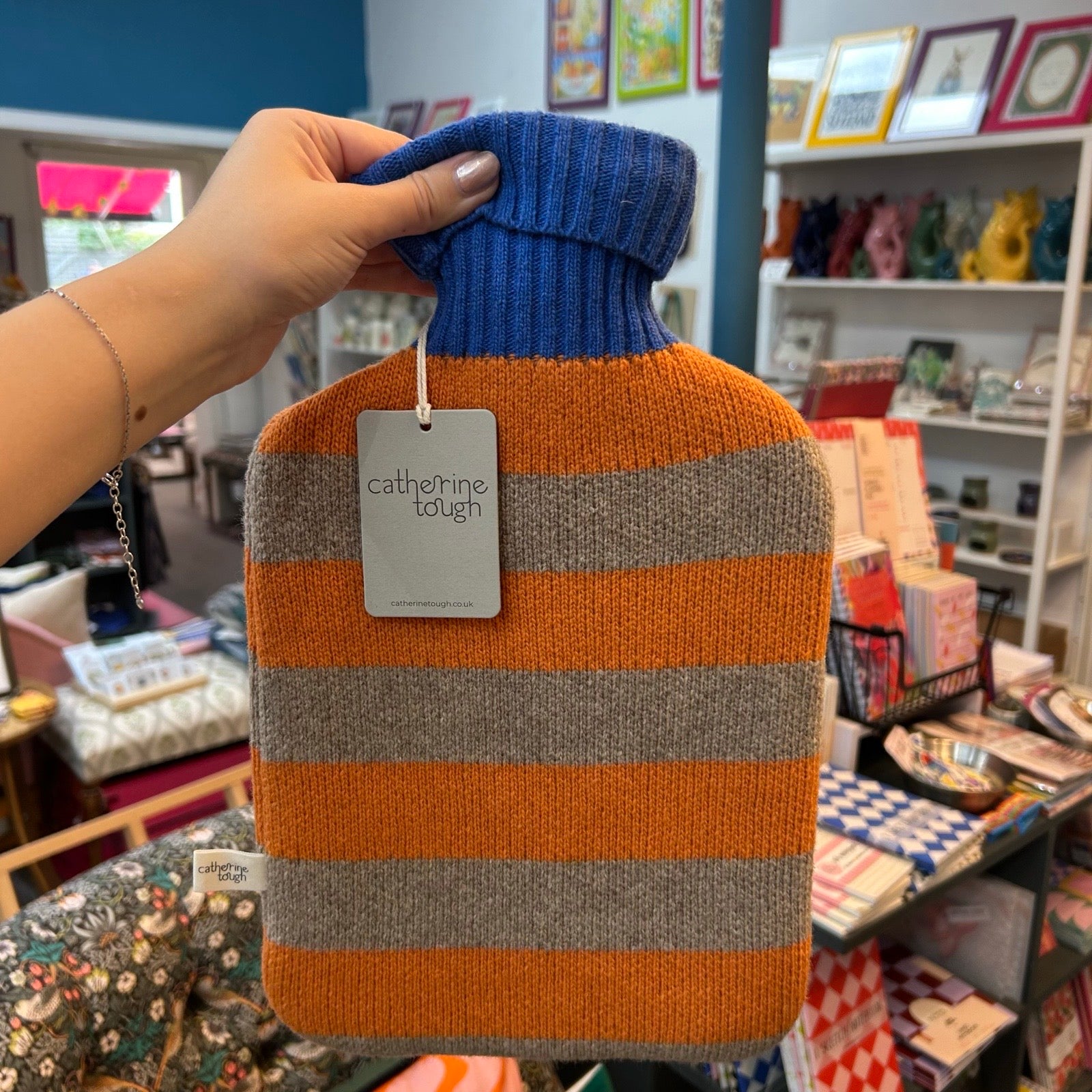 A hand holds a knitted hot water bottle cover with horizontal stripes in orange, grey, and blue neck. The tag on the product reads 'catherine tough'.
