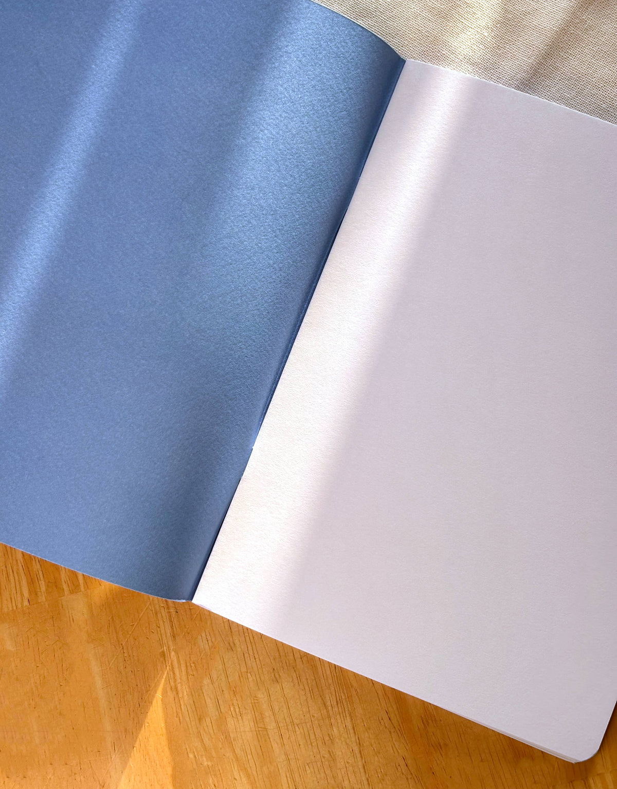 A close-up of an open notebook or book showing a textured blue left page and a plain white right page, resting on a wooden surface with a beige fabric surface above it.