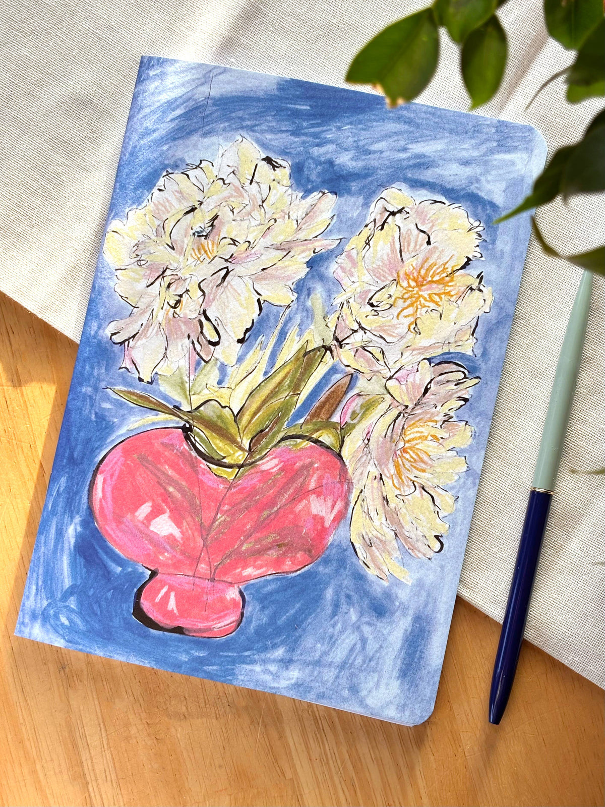 A square art print or notebook cover featuring a painted floral design with white and pale yellow flowers in a pink vase on a blue background, styled on a wooden surface with a pen and fabric.