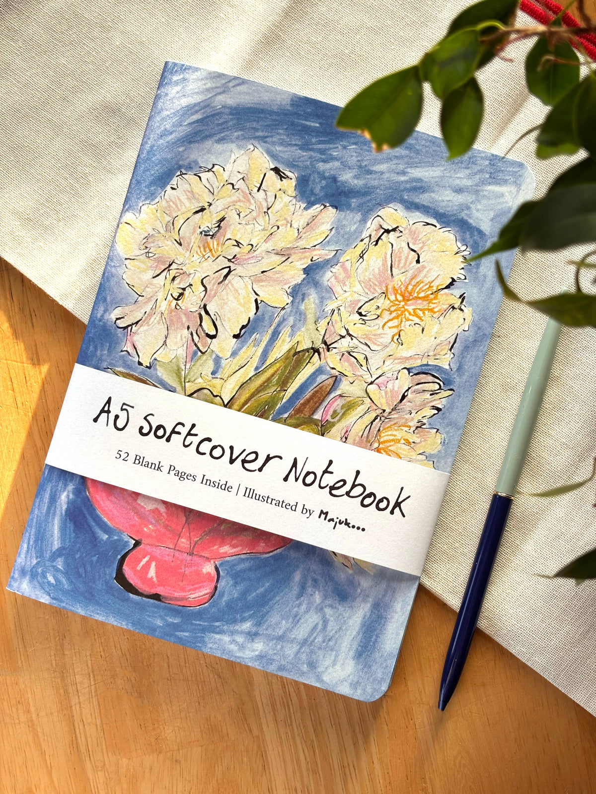 A notebook with an illustrated floral cover lies on a wooden surface next to a beige fabric cloth, a green pen, and plant leaves. The notebook has a white paper band reading 'A5 Softcover Notebook' and '52 Blank Pages Inside | Illustrated by Majukoo'.