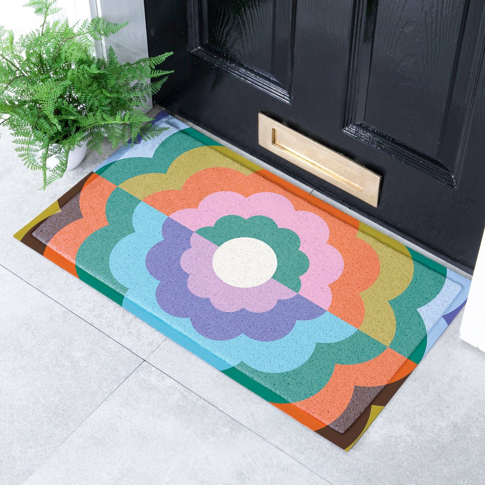 A rectangular doormat with a retro floral and geometric pattern in multiple colours, placed in front of a black front door on a light grey tiled floor next to a potted green plant.