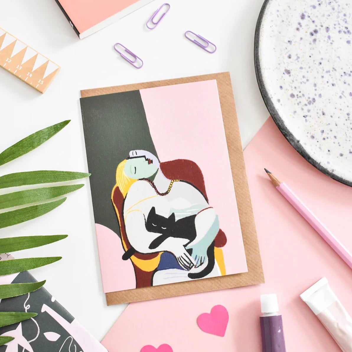Greeting card with an illustration of a woman and a cat on a pink background, surrounded by stationery items.