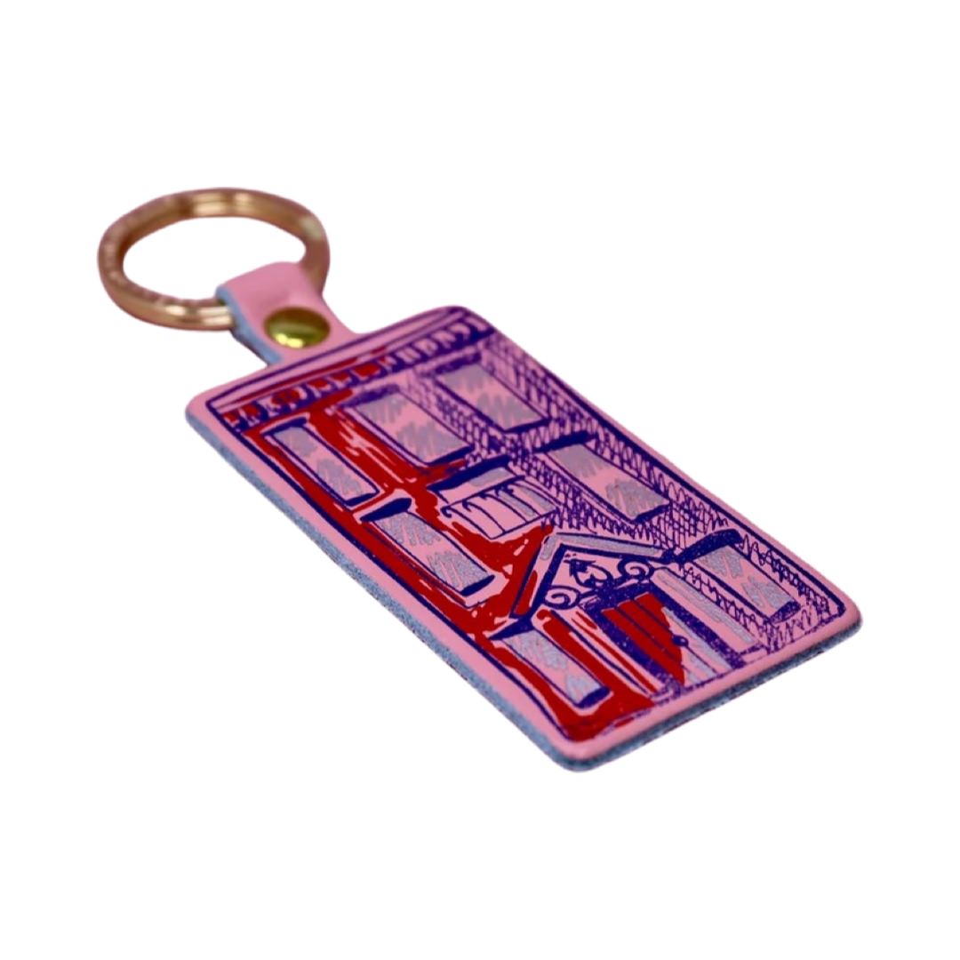 A pink rectangular leather keyring with an illustration of an apartment block in purple and a gold-coloured keyring. 