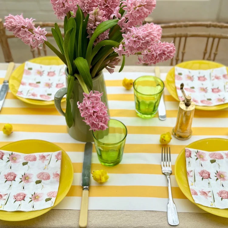 Dining table setting with pink flowers, green glasses, and yellow plates on a striped tablecloth.