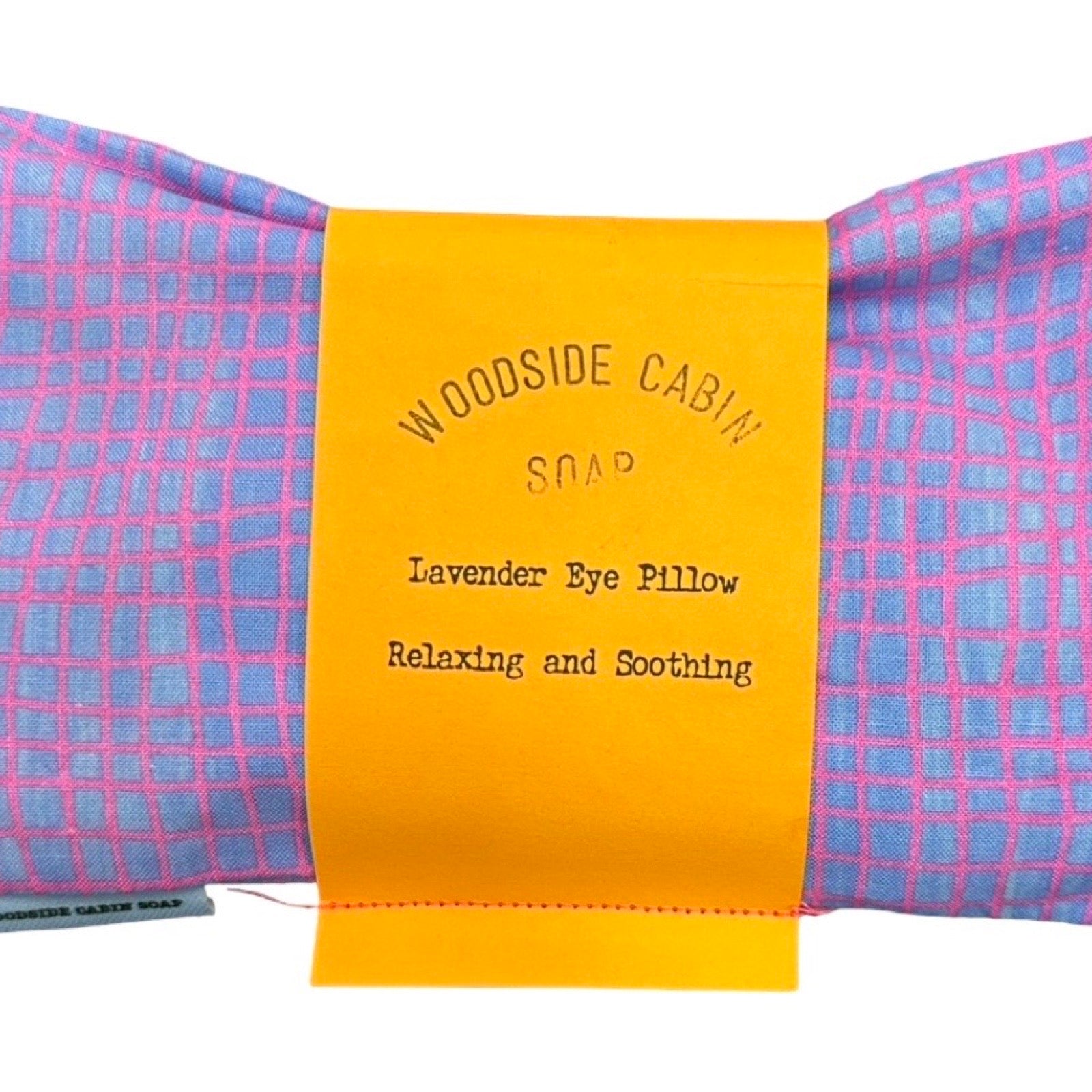 A lavender eye pillow by Woodside Cabin Soap, featuring a pink and blue checkered fabric body with a bright yellow paper label wrapped around it.