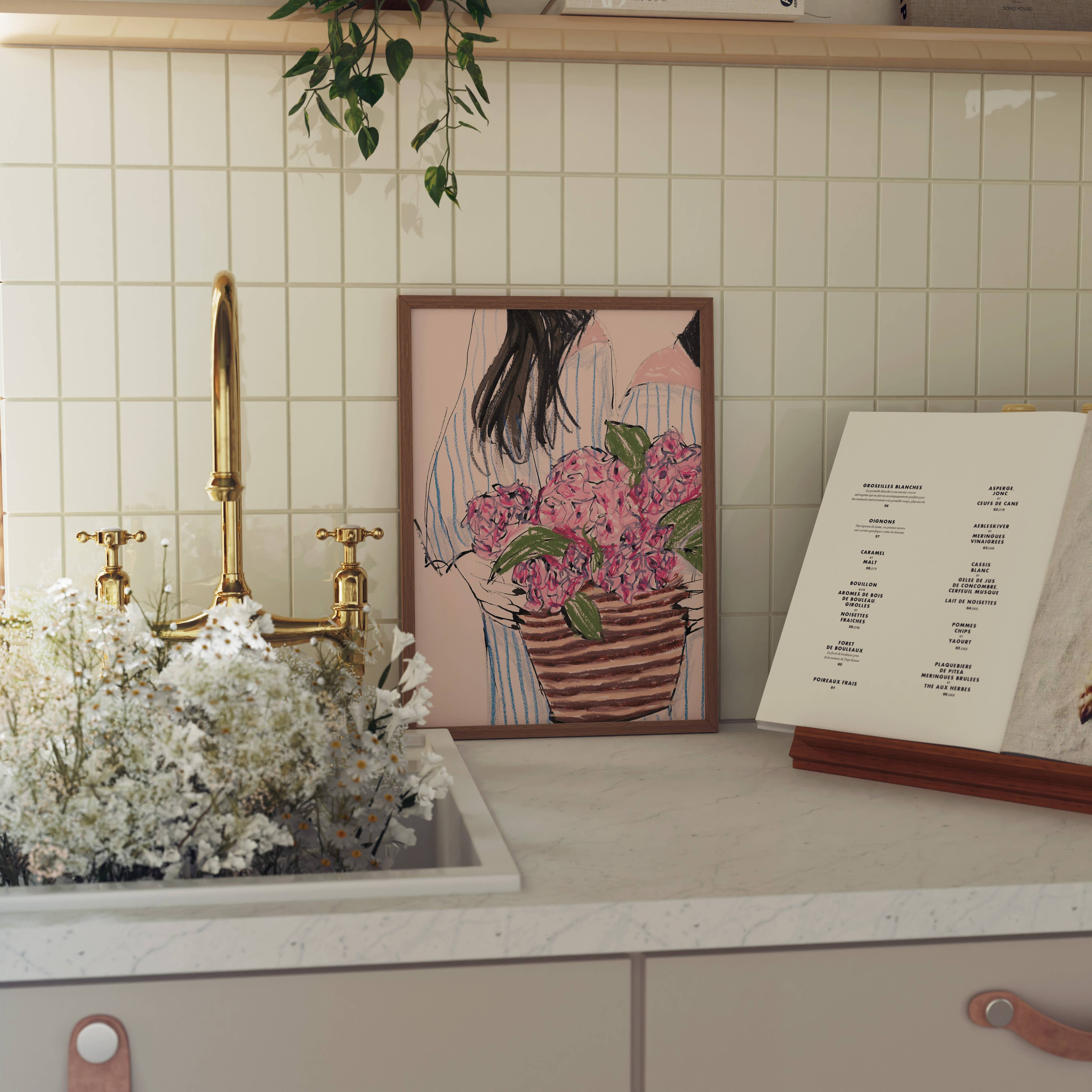 A framed art print depicting a woman holding pink flowers, styled on a kitchen counter next to a gold faucet and white flowers, with a menu card on a stand.