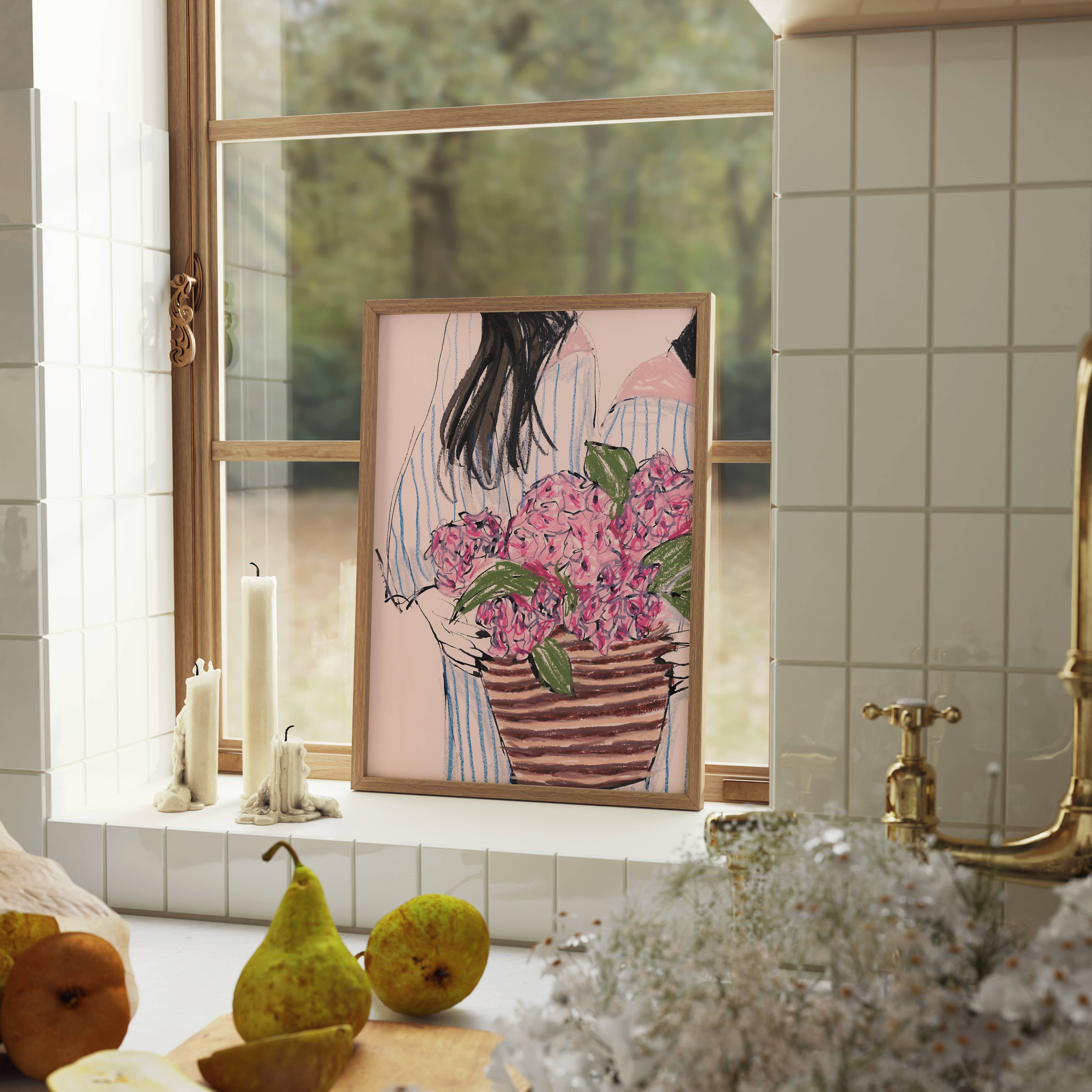 A framed art print featuring an illustration of a woman holding a large pink flower bouquet, displayed on a kitchen windowsill with candles, fruit, and a brass faucet in the background.