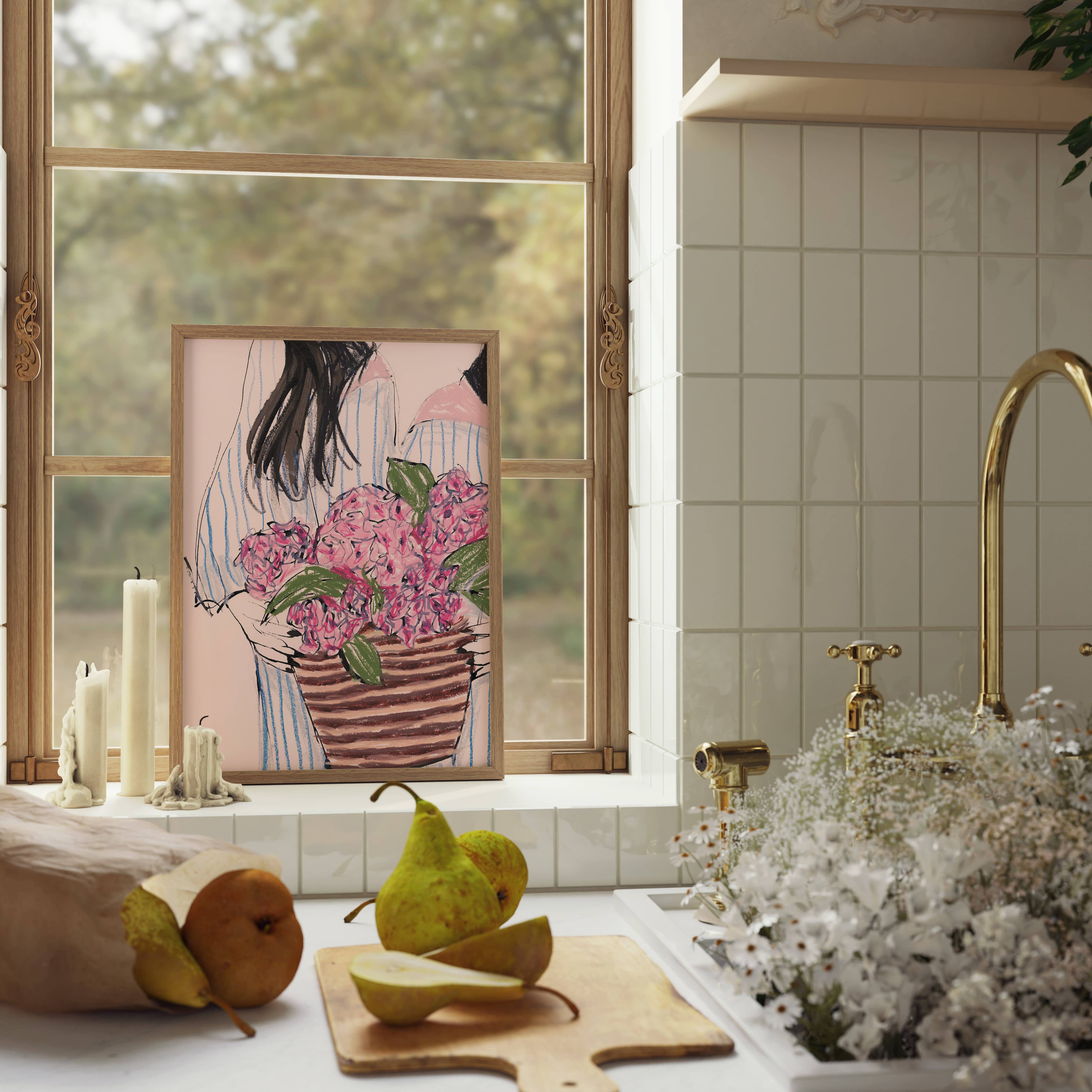 A framed art print featuring an illustration of a woman holding a basket of pink flowers, styled on a kitchen windowsill next to candles, pears, and a sink with white flowers.