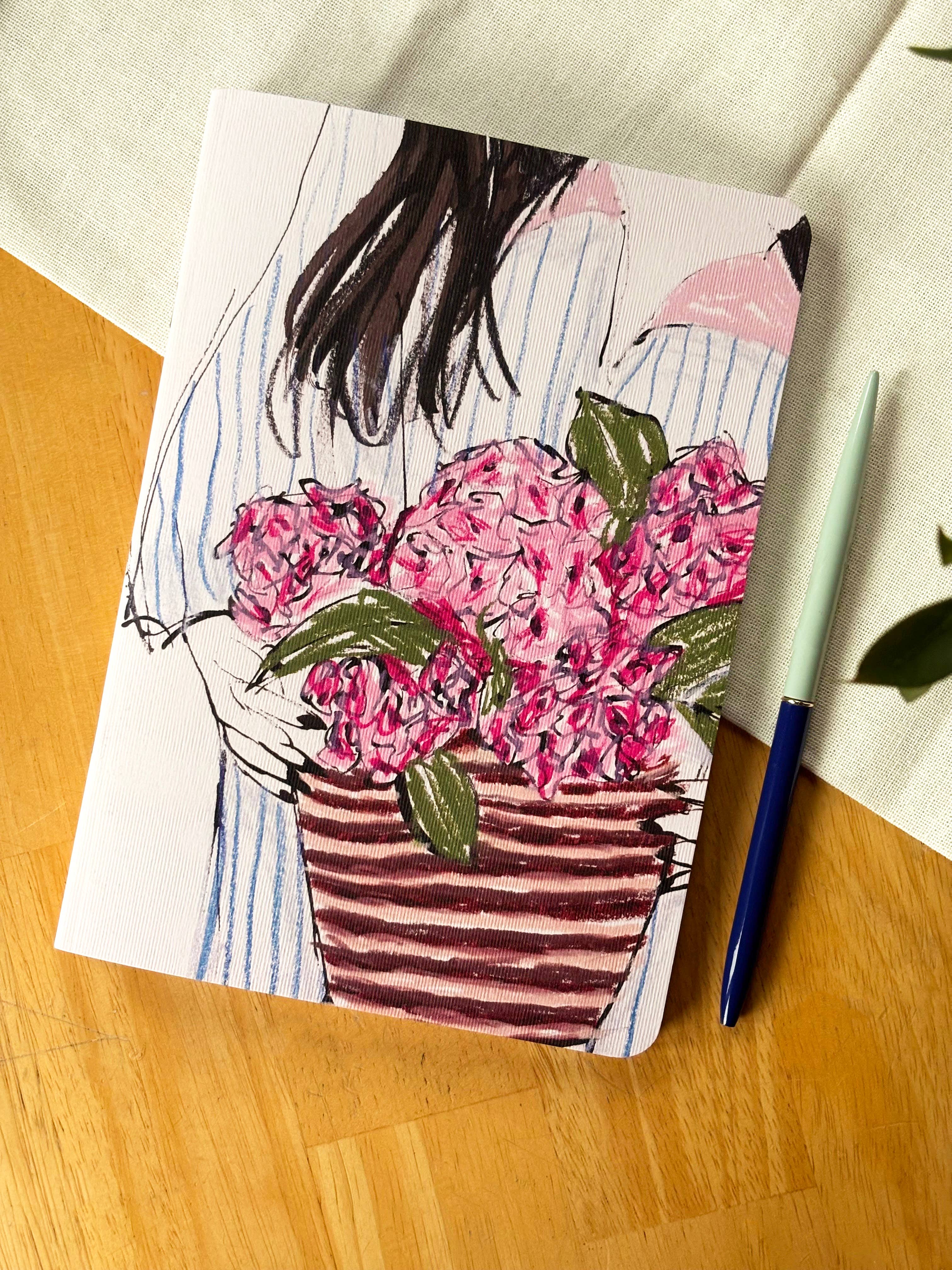 A square art print or notebook cover featuring a hand-drawn illustration of a woman holding a large pink flower bouquet in a striped basket, placed on a wooden surface next to a pen and white fabric.
