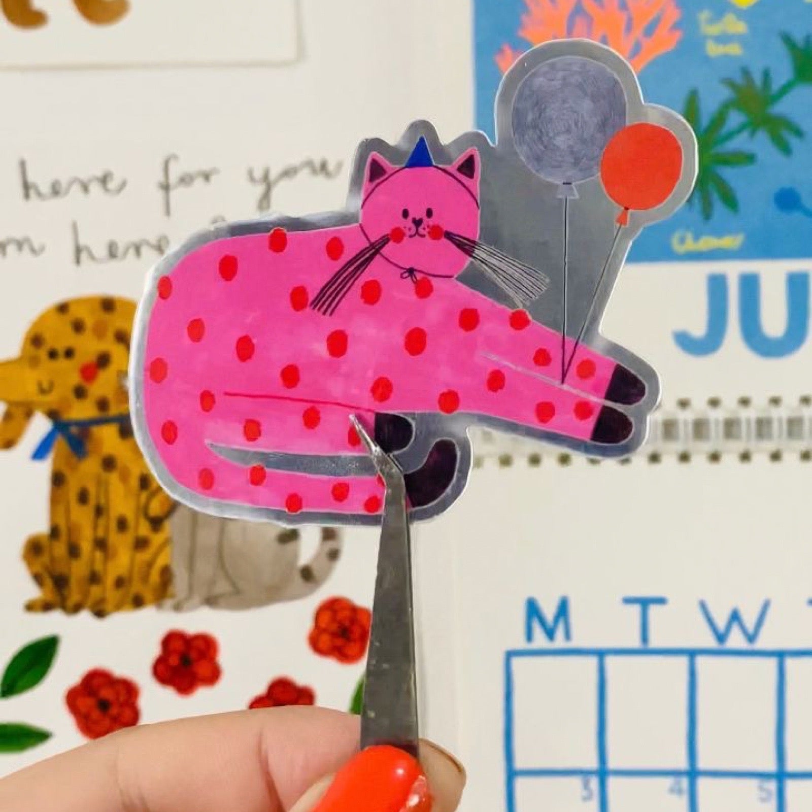 Pink polka dot cat sticker held by a hand with orange nail polish and tweezers, against a colourful background.