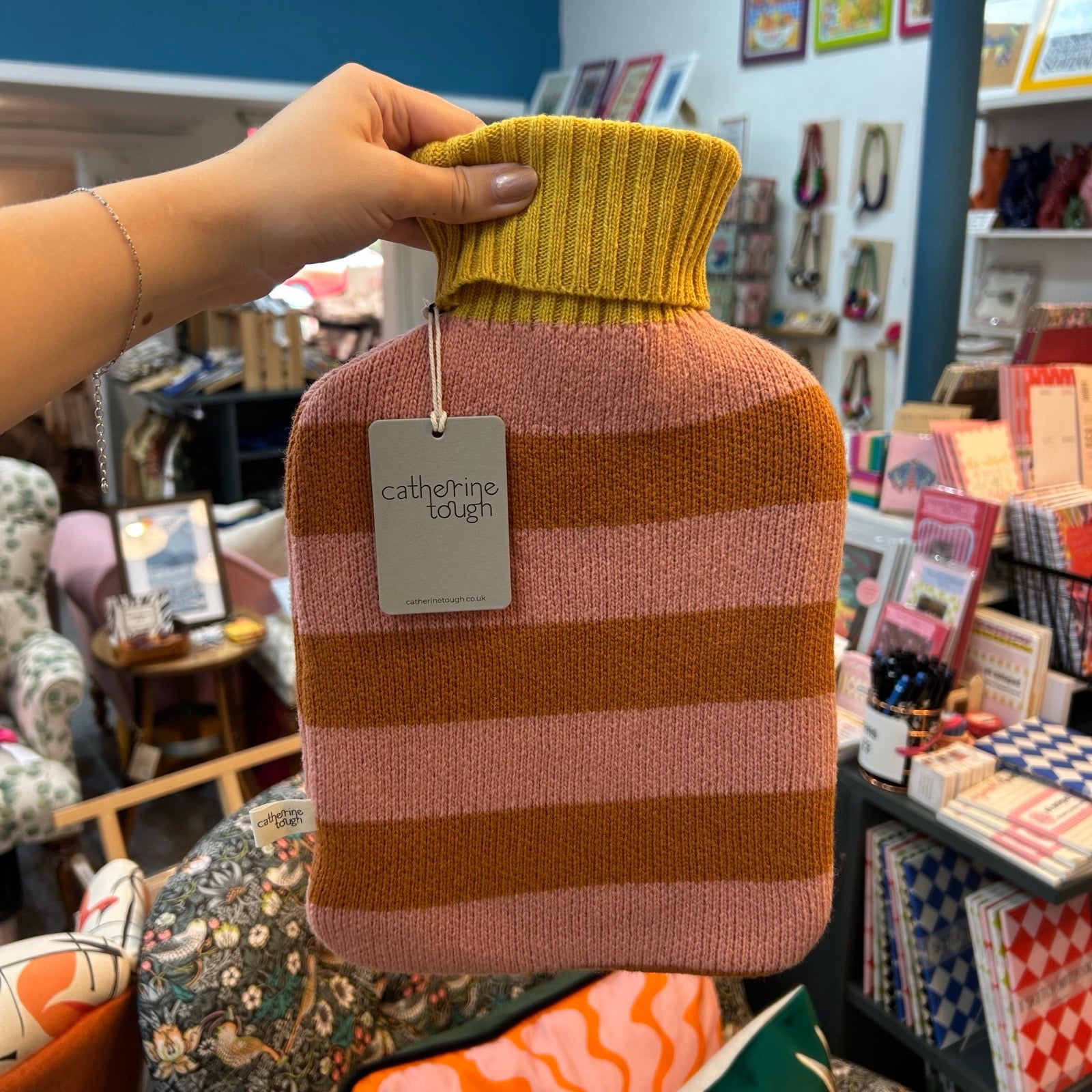A hand holds a hot water bottle covered in a knitted sweater-style sleeve. The sleeve has horizontal stripes in mustard yellow, dusty pink, and rust orange, with a grey tag reading 'catherine tough'.