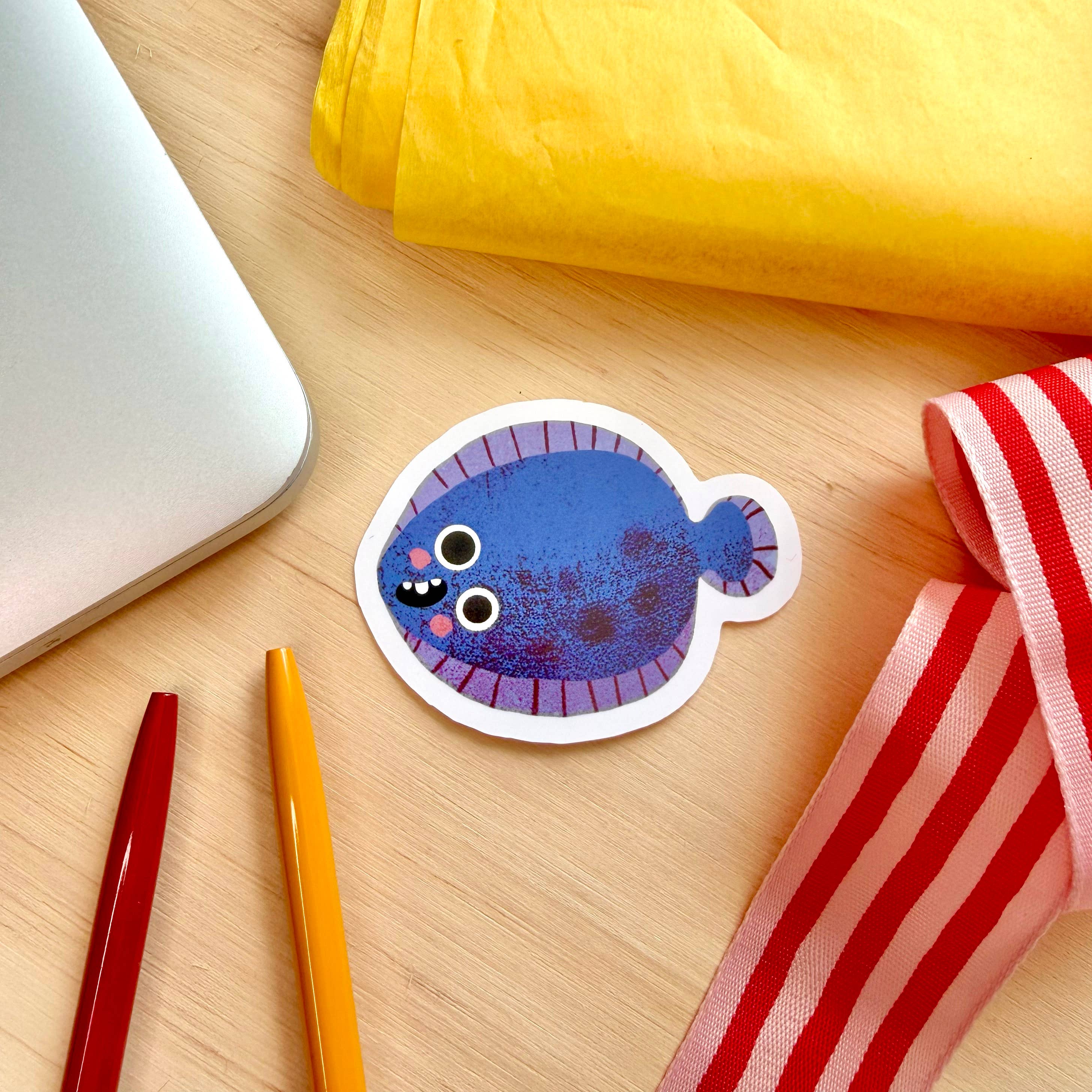 A die-cut sticker of a blue flatfish with a purple striped border, placed on a light wood desk alongside a silver laptop, yellow paper, red and orange pens, and a red-and-white striped ribbon.