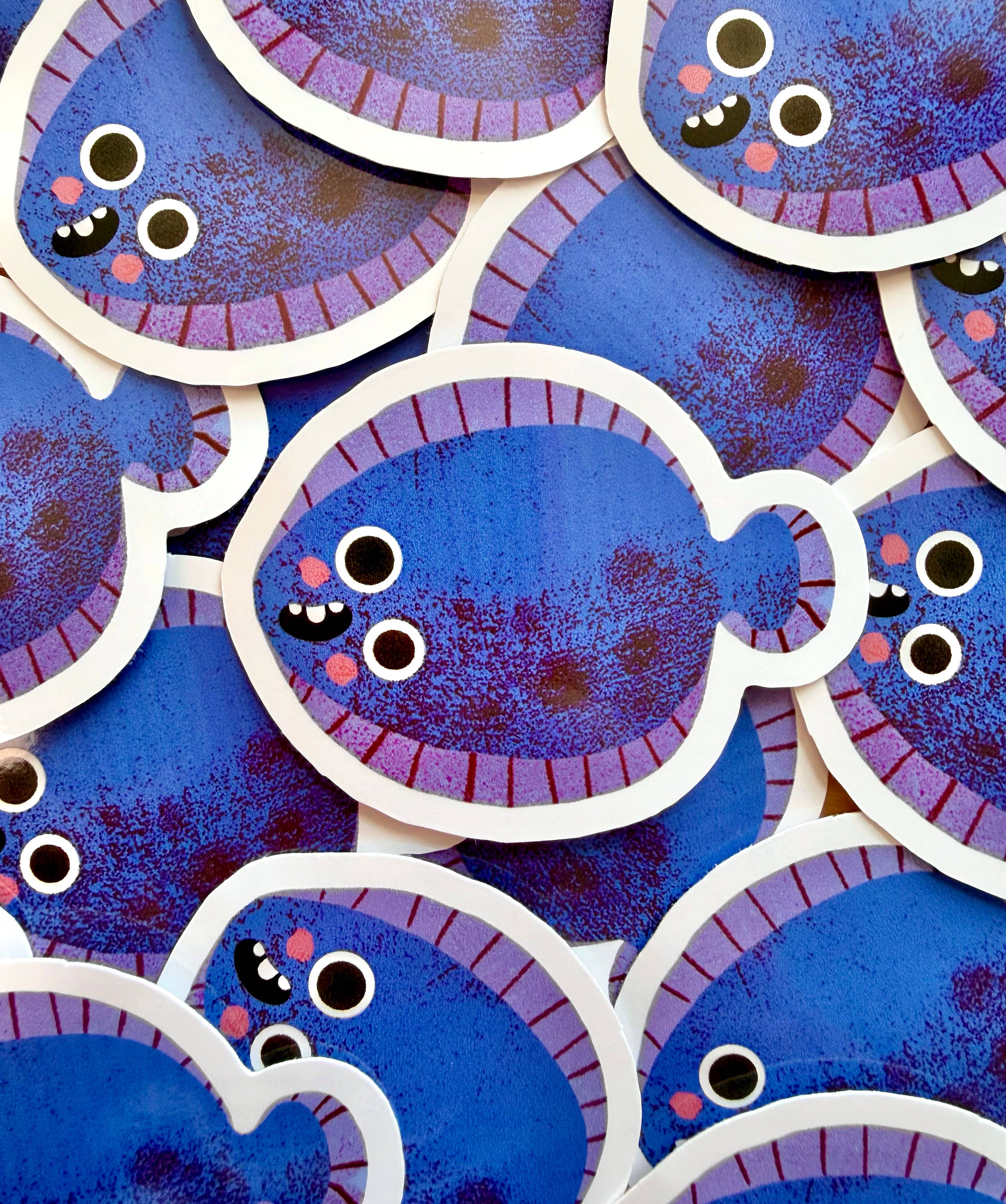 A close-up of multiple overlapping fish-shaped stickers or decals. Each fish has a textured blue body, a purple scalloped border, and a white outline, with simple cartoon eyes and small pink cheeks.