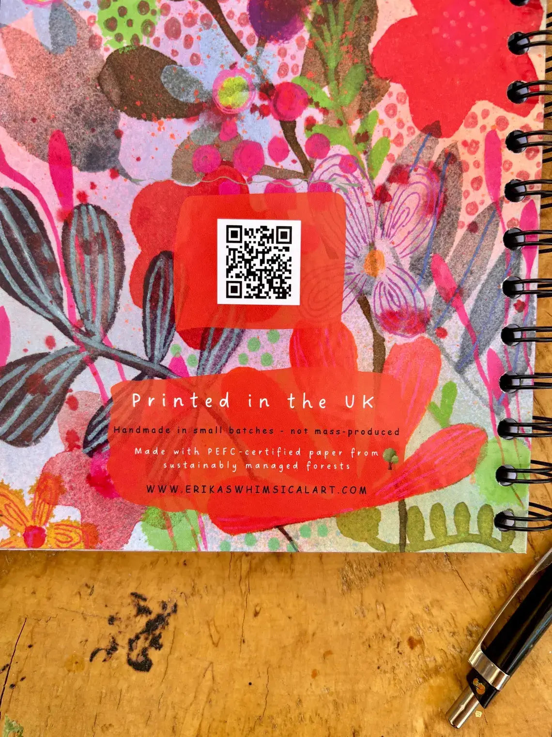 A spiral-bound notebook with a vibrant, multicolored floral watercolor cover design, resting on a wooden tabletop next to a black pen. The cover features a QR code and 'Printed in the UK' text.