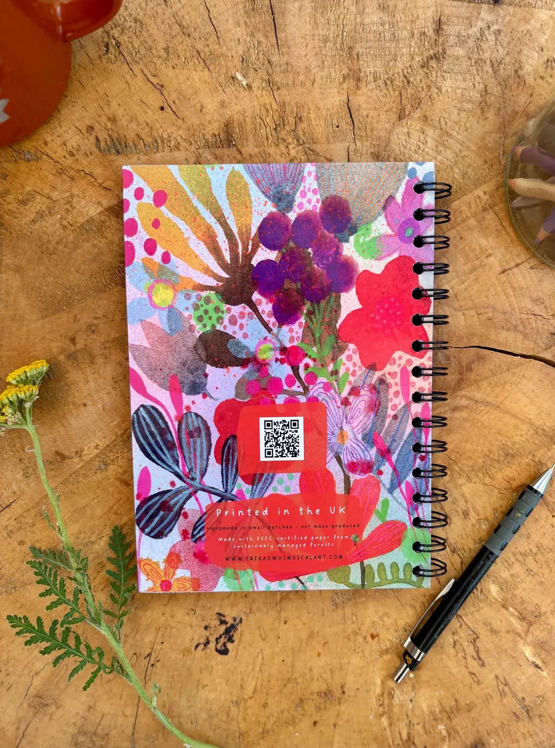 A spiral-bound notebook with a colourful floral and abstract botanical print lies on a rustic wooden tabletop. A small red sticker with a QR code is attached near the bottom, with partial text reading 'Printed in the UK'.