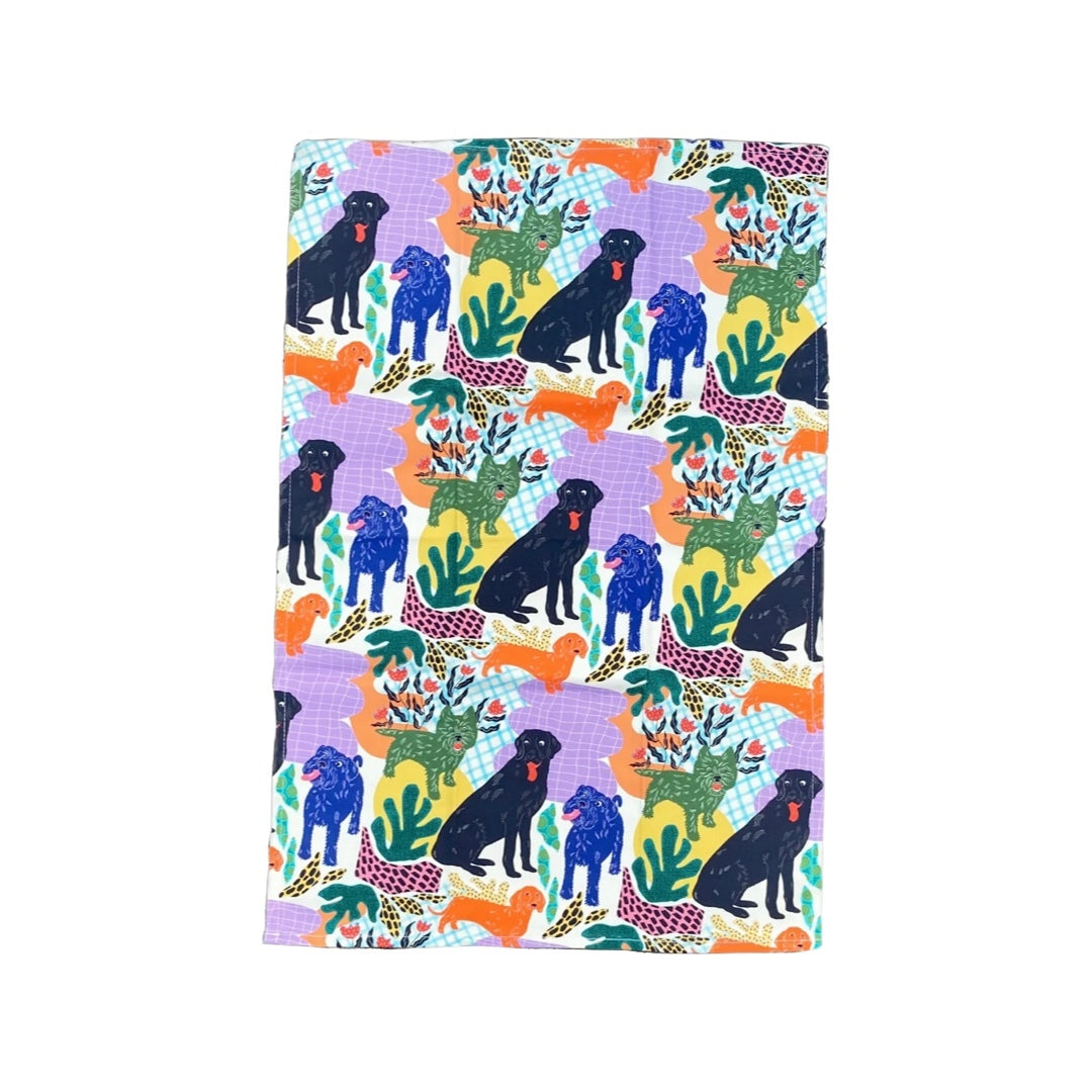 Dog lovers tea towel featuring black labradors, orange dachshunds and green scottie dogs with green leaves and a lilac background.