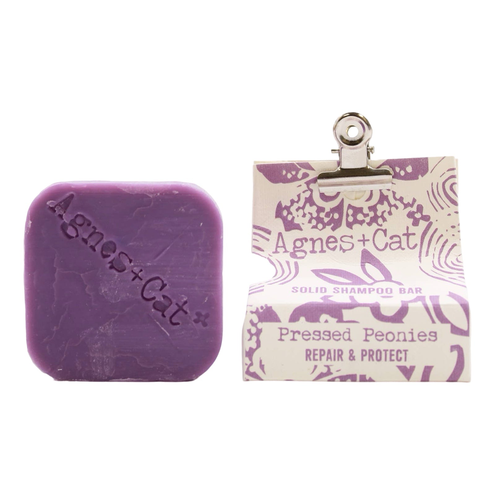 A square-shaped purple, pressed peonies scented dry shampoo bar and a white and purple packet held closed with a bulldog clip.