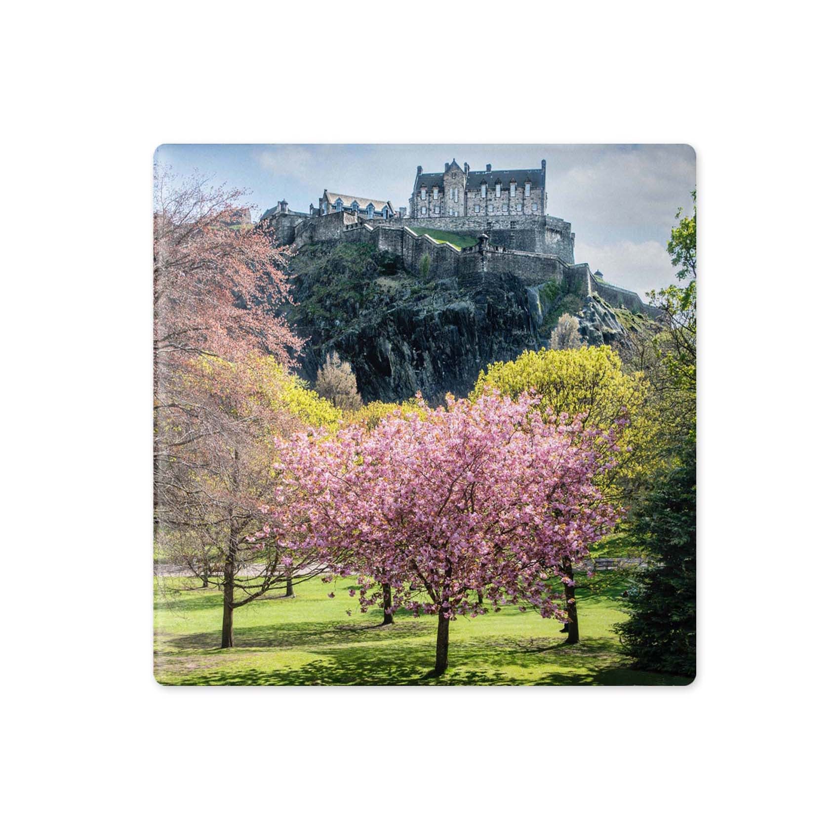 A square coaster featuring a scenic photograph of Edinburgh Castle on a rocky hill, with blooming pink and yellow trees in the foreground and a grassy park below.