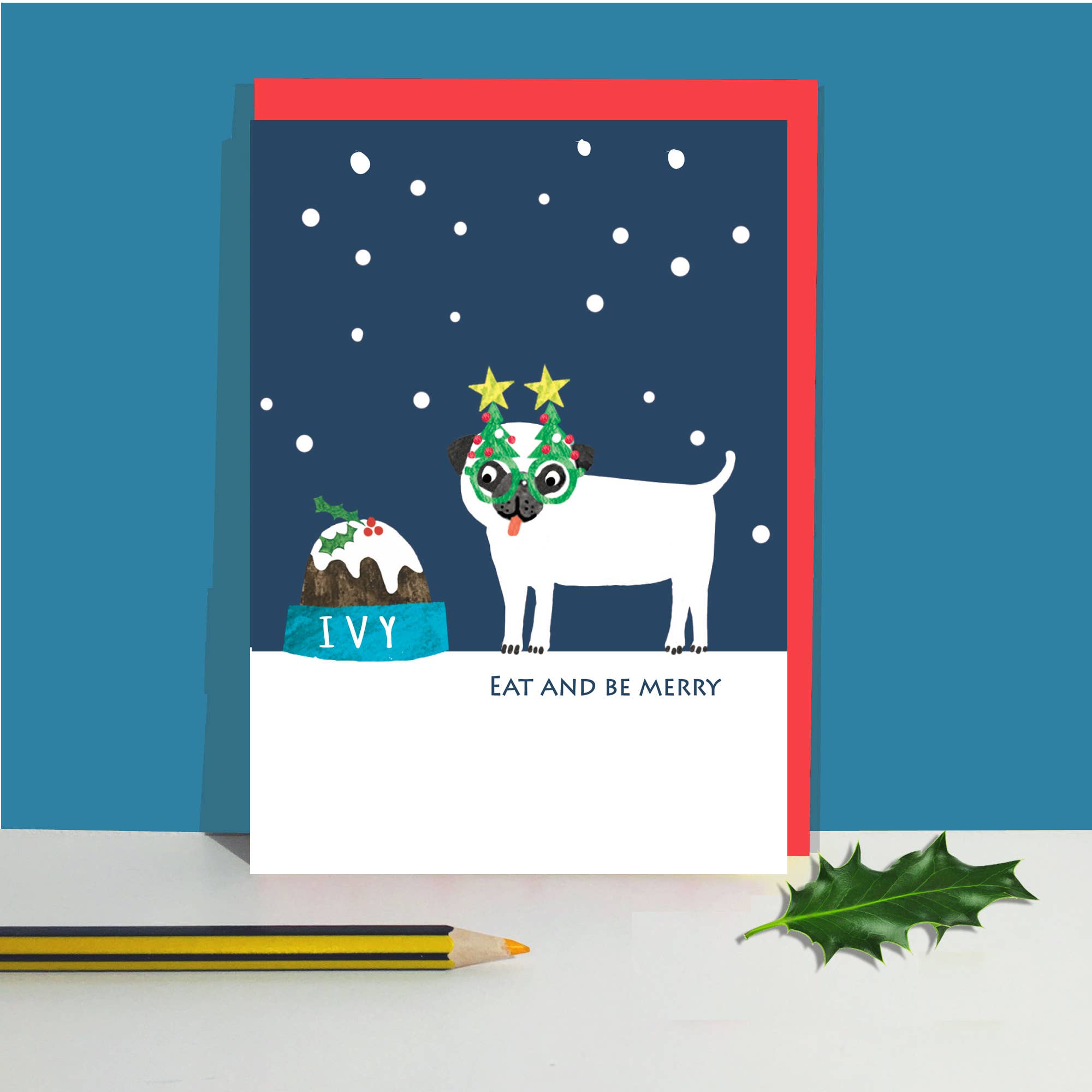 A Christmas-themed greeting card featuring a white pug wearing green Christmas glasses and a star crown, standing next to a decorated cake with the name 'IVY'. The card has a red envelope, is displayed on a white surface with a green holly leaf and a yellow pencil, set against a blue background.