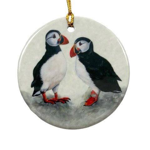 A circular ceramic decoration featuring an illustration of two standing puffins. The decoration is printed on both sides and has a gold cord for hanging.