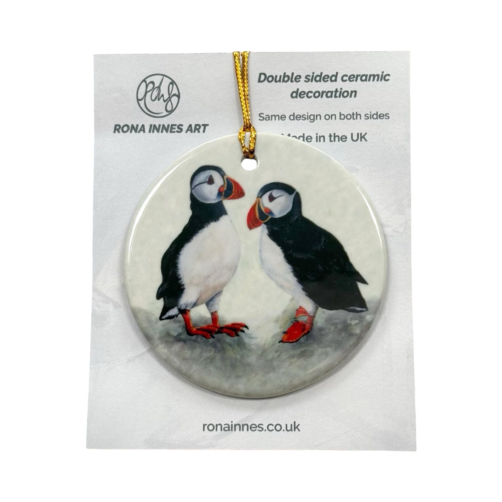 A circular ceramic decoration featuring an illustration of two standing puffins. The decoration is printed on both sides and has a gold cord for hanging.