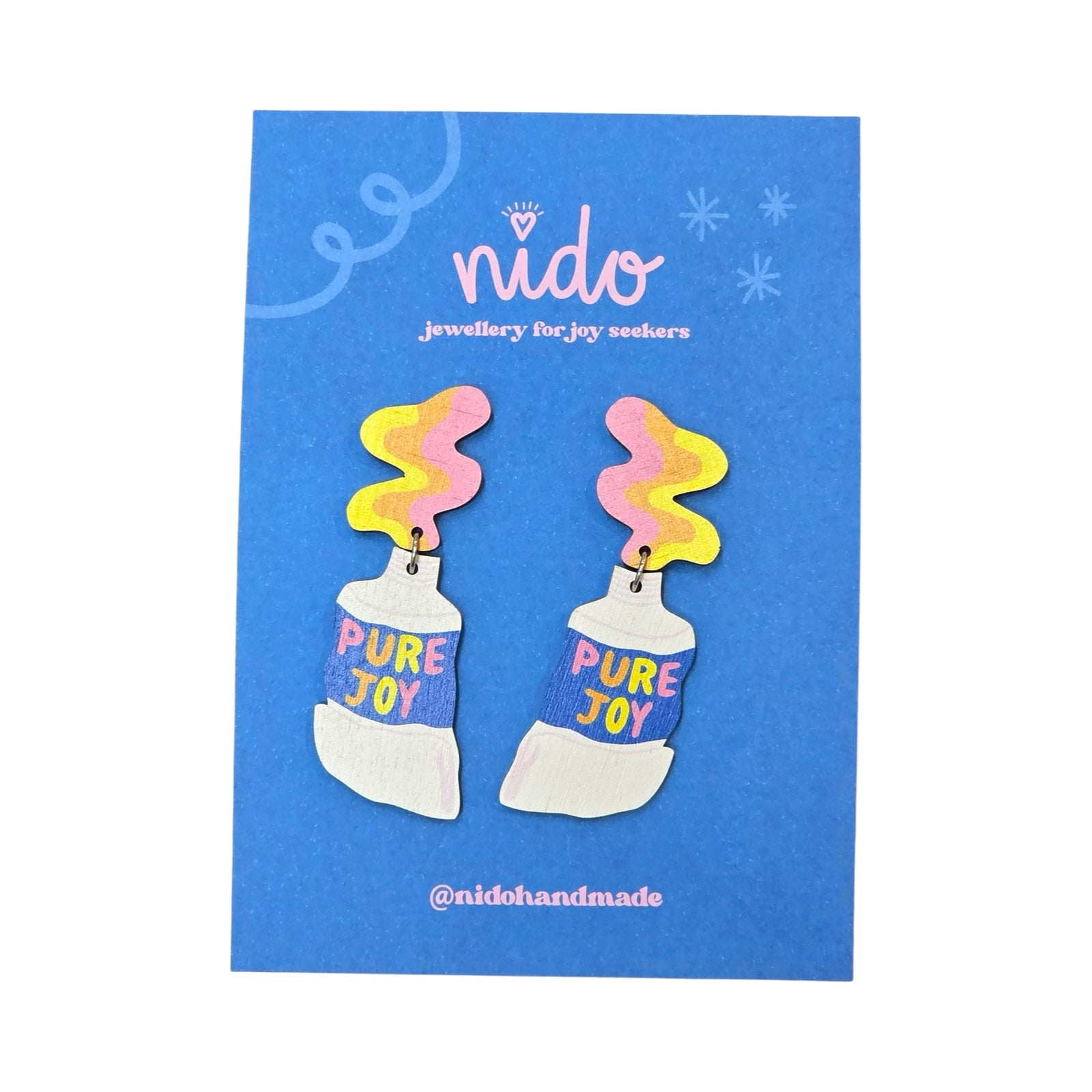 These wooden paint tube earrings feature yellow, orange and pink paint being squeezed out of a white tube of paint with the multicoloured text 'Pure Joy' in the centre of the tubes, presented on a branded blue backing card.