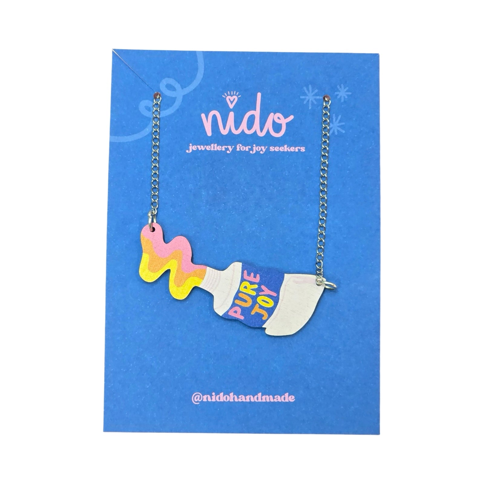 This wooden paint tube necklace features yellow, orange and pink paint being squeezed out of a white tube of paint with the multicoloured text 'Pure Joy' in the centre of the tubes, presented on a branded blue backing card.