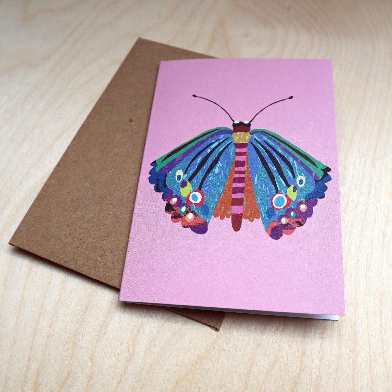 A pink greeting card featuring a colorful painted butterfly, shown with a brown kraft envelope on a light wood surface.