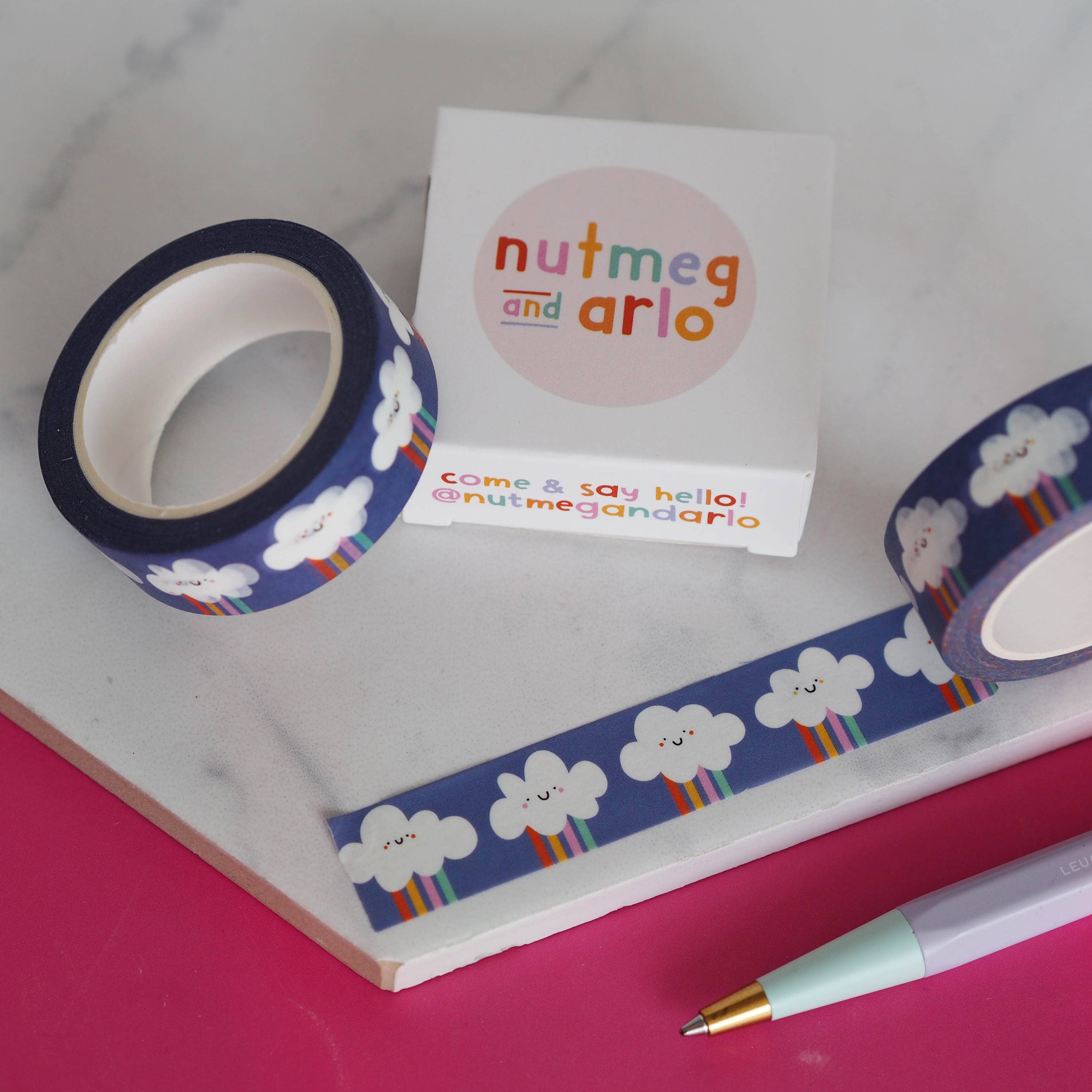 Washi tape with cloud design, a notebook with cloud-themed border, and a pen on a pink surface.