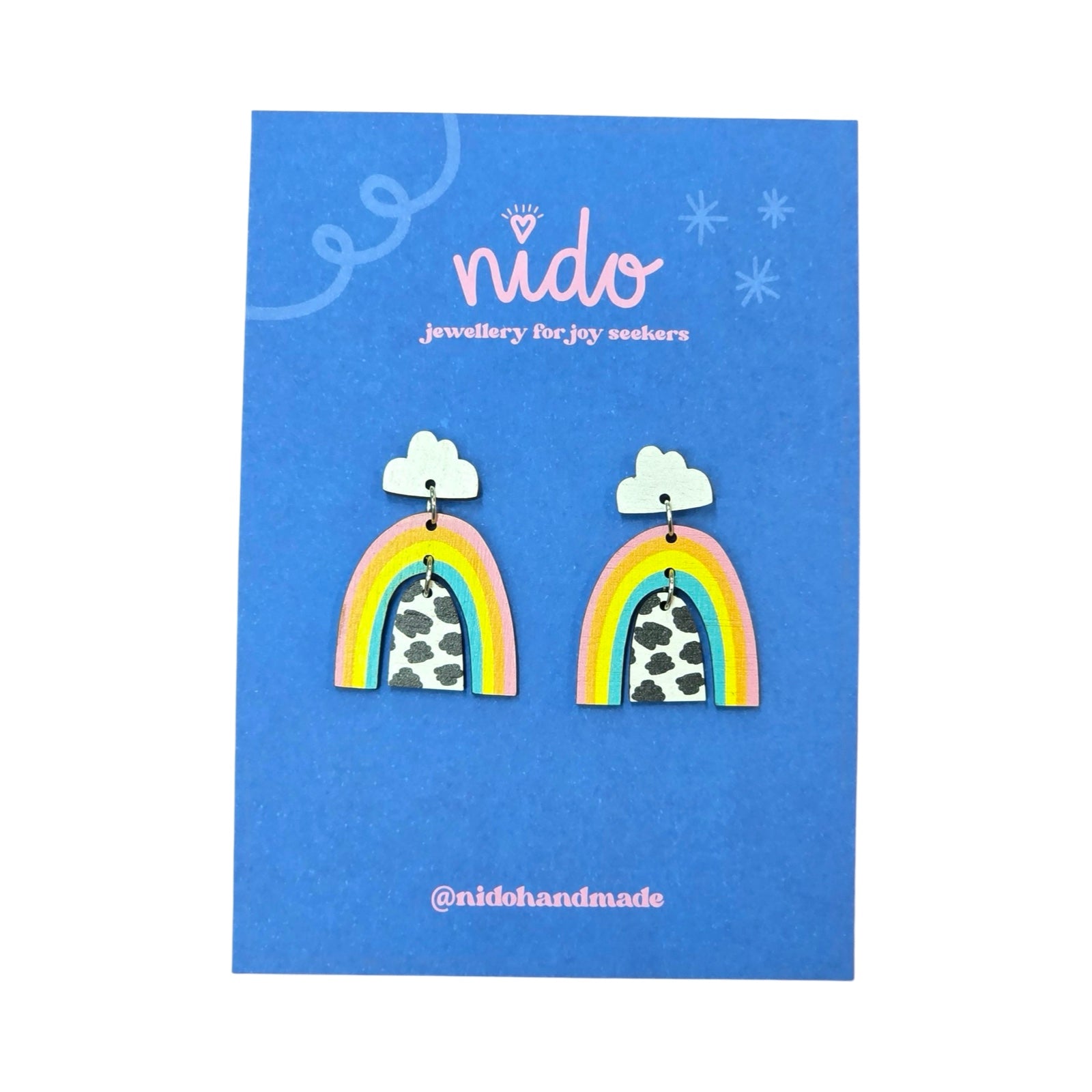 These wooden earrings feature a white cloud stud with a pink, orange, yellow and teal rainbow hanging underneath and a black and white pebble effect arc hanging inside, presented on a branded blue backing card.