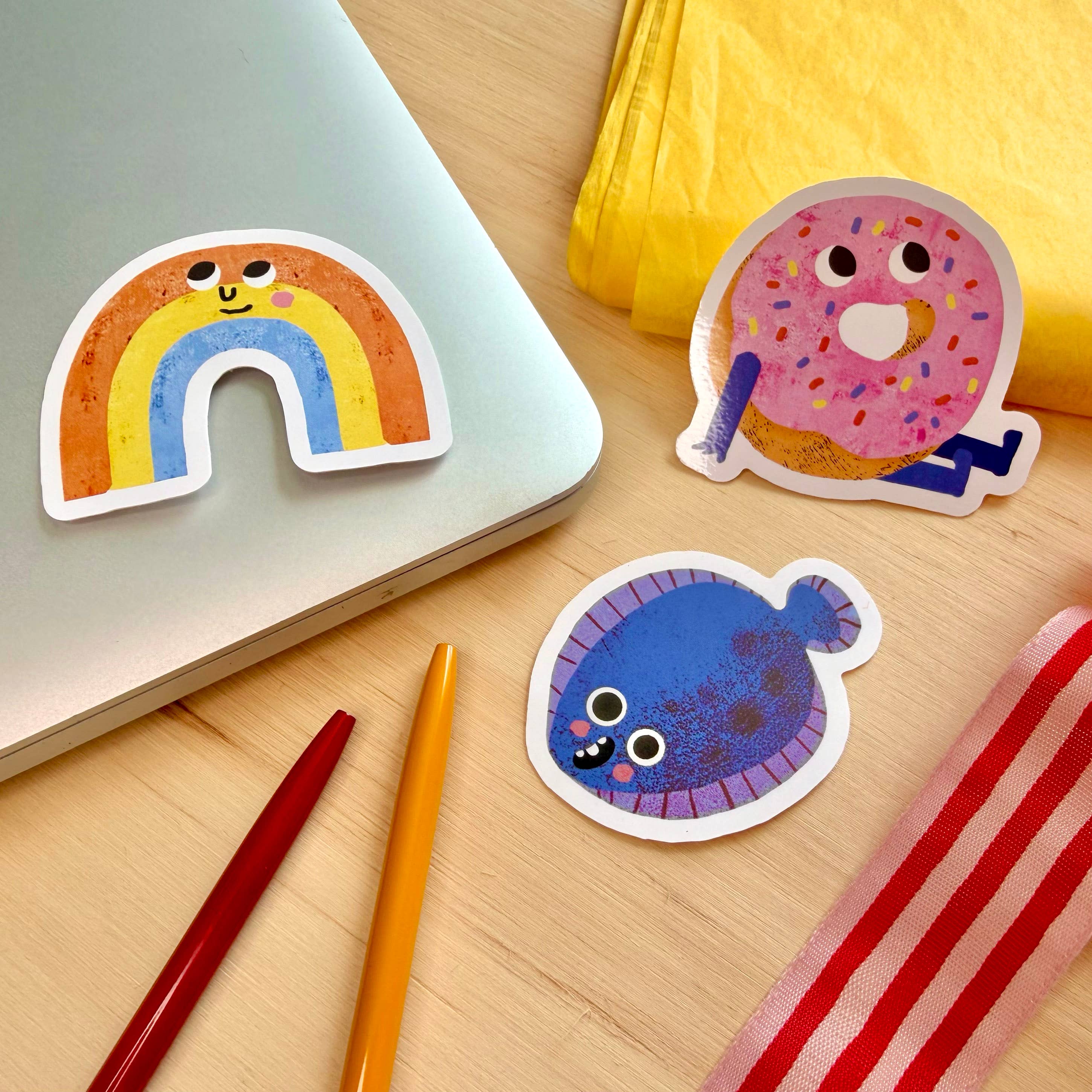 Three colourful cartoon stickers on a light wood desk: a rainbow sticker on a laptop, a pink donut sticker, and a blue fish sticker, with pens, ribbon, and a yellow tissue paper.