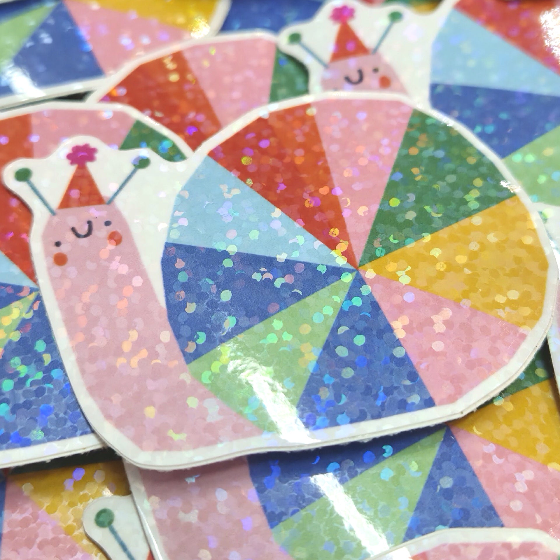 A close-up of colorful glitter snail stickers arranged on top of each other. The main sticker shows a pink snail wearing a party hat, with a segmented shell made of pastel rainbow glitter triangles.