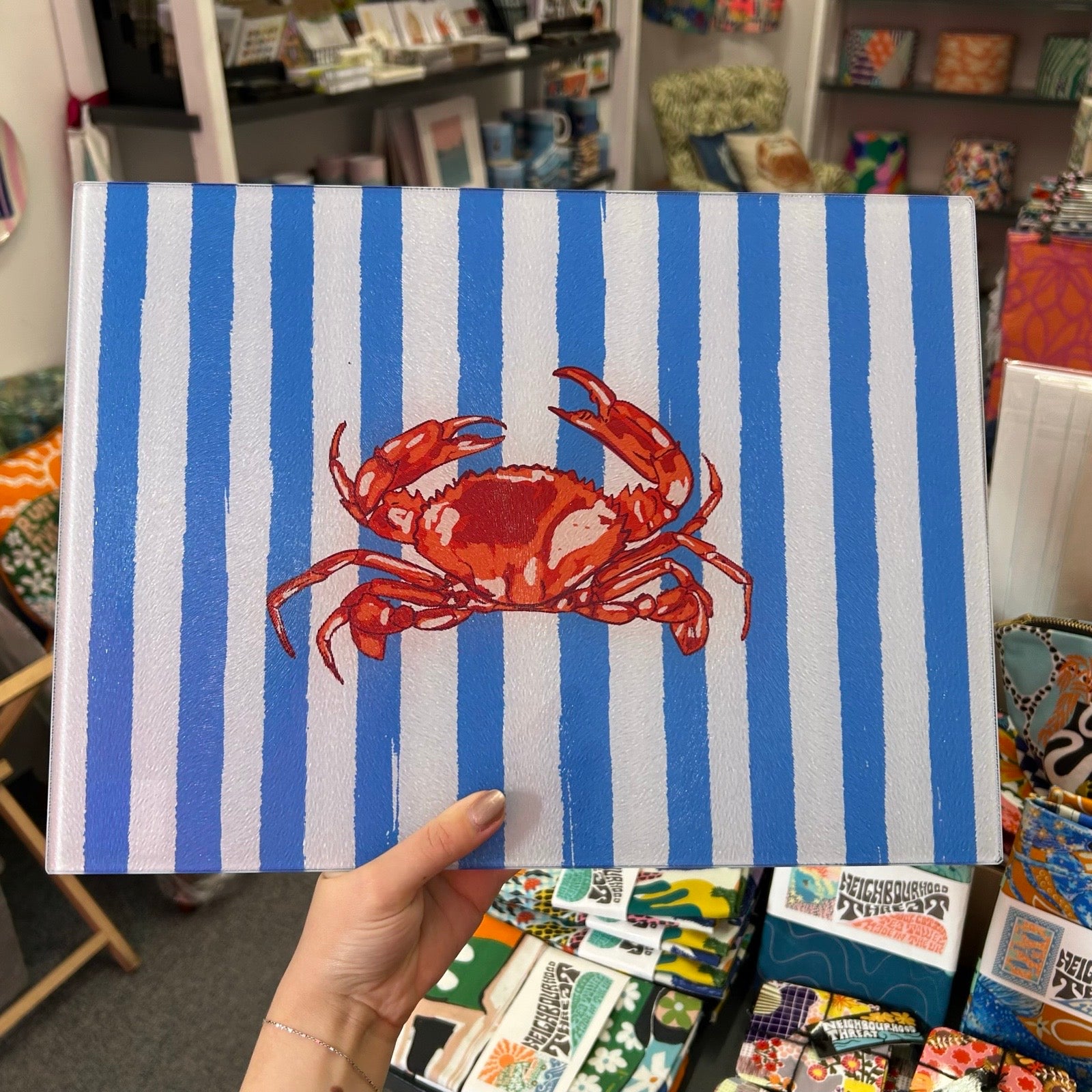 A hand holds a chopping board featuring a red crab on a blue and white striped background, with a retail store interior visible in the blurred background.