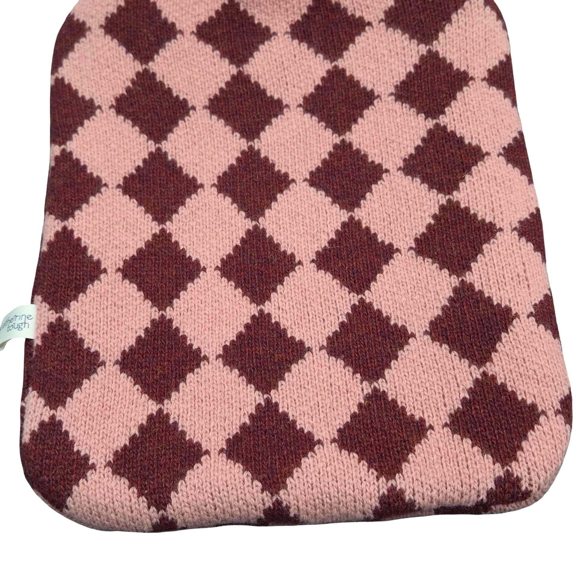 A knit hot water bottle cover featuring a classic argyle pattern in dusty pink and dark brown, with a small white fabric tag on the left edge.