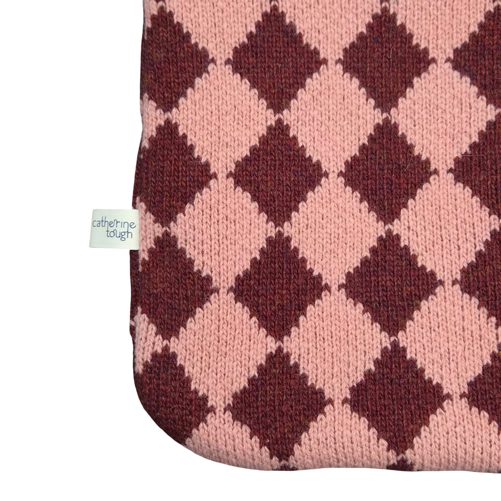Close-up of a knitted textile with an argyle diamond pattern in dusty pink and dark burgundy, showing a small white tag that reads 'catherine tough' on a plain white background.