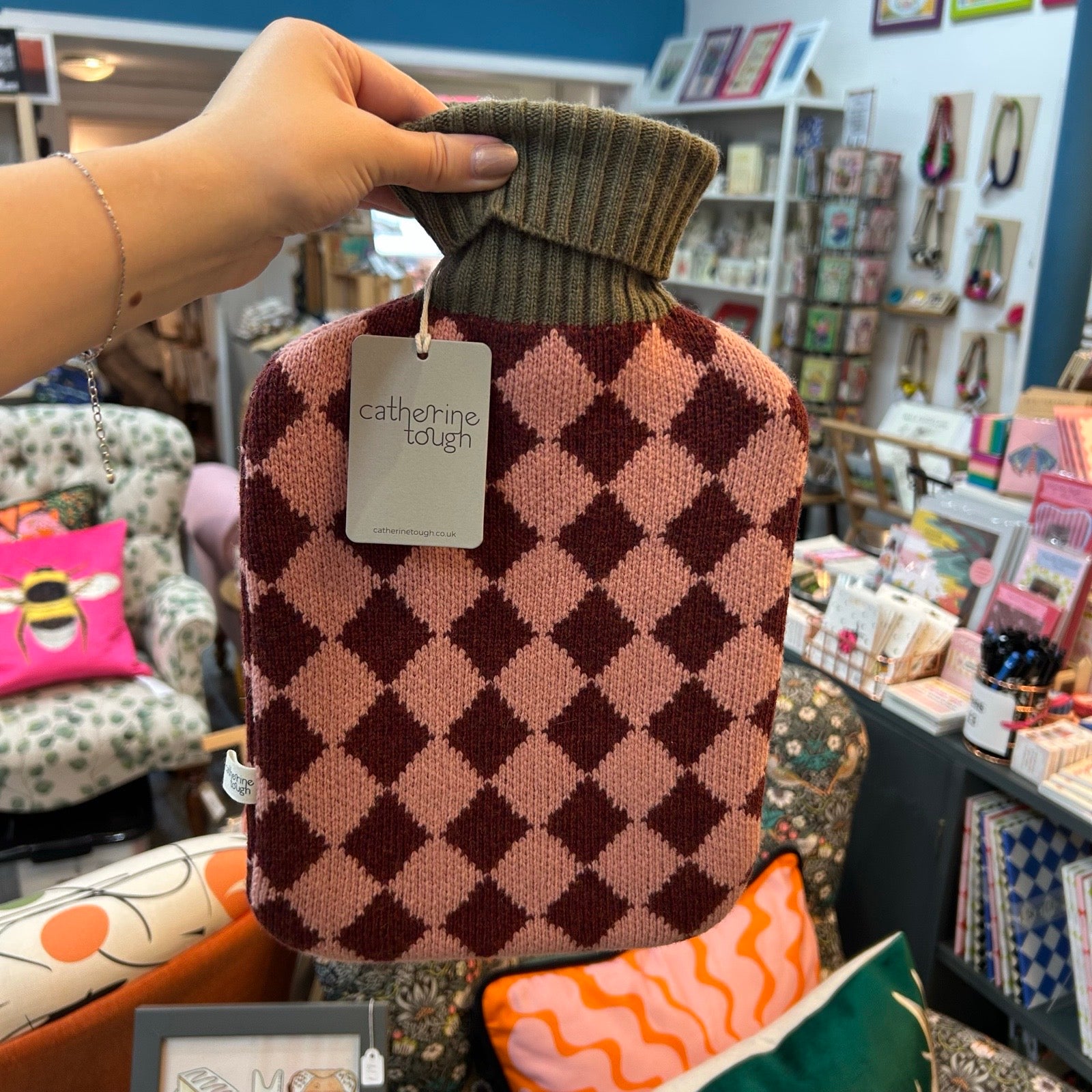 A hand holds a hot water bottle cover with a pink and burgundy argyle pattern and an olive green ribbed neck. A tag reading 'catherine tough' is attached.
