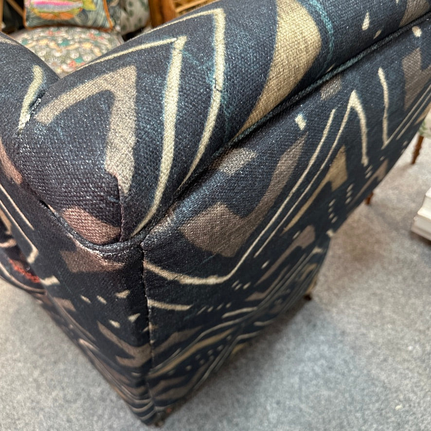 Close-up of an upholstered chair with a dark abstract patterned fabric in charcoal, beige, and taupe tones, shown in an indoor setting on a grey carpet.