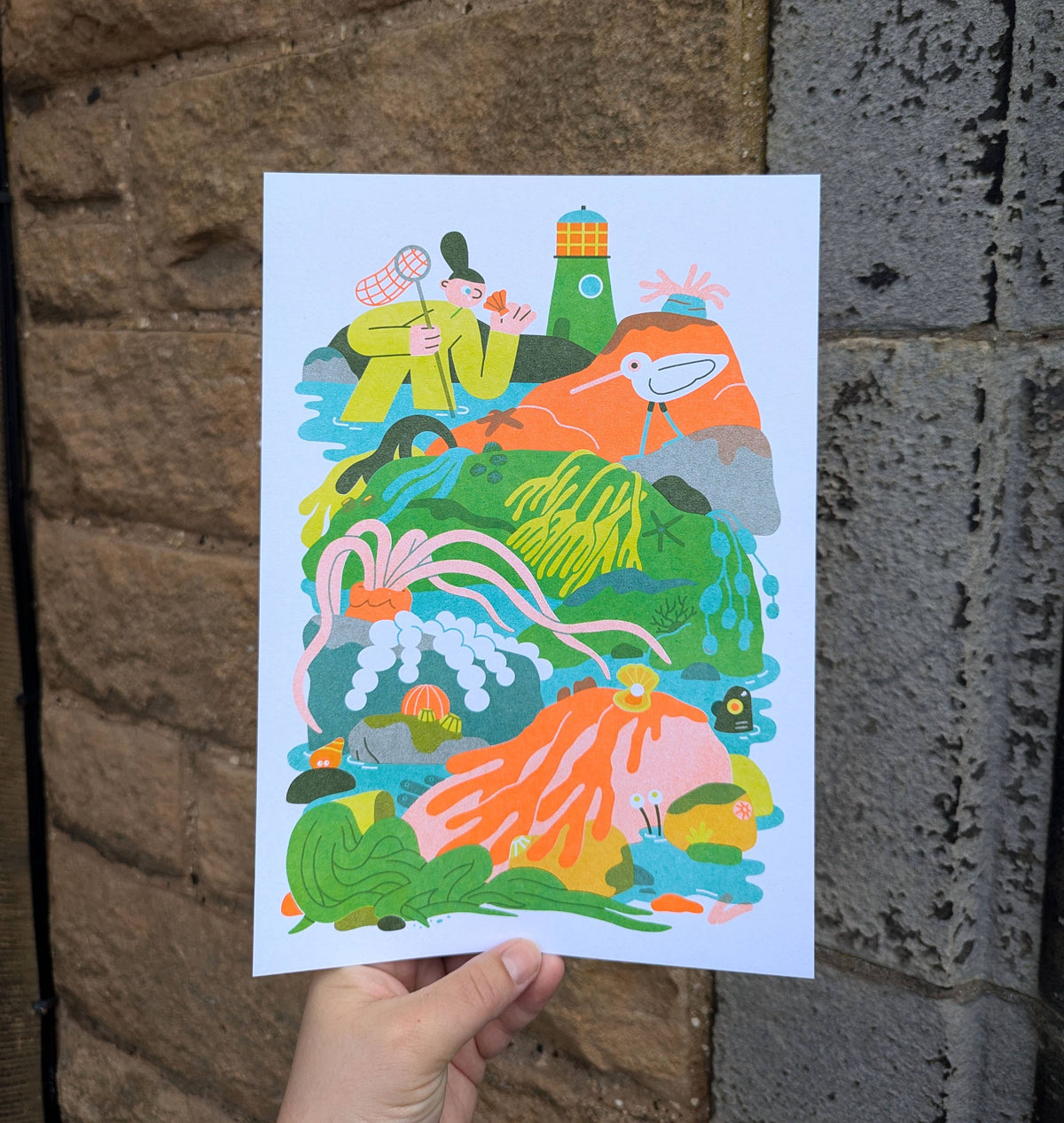 A hand holds a white art print featuring a colourful whimsical illustration of a person in a yellow top next to a lighthouse and a bird, set against a background of a brown brick wall and a grey concrete wall.