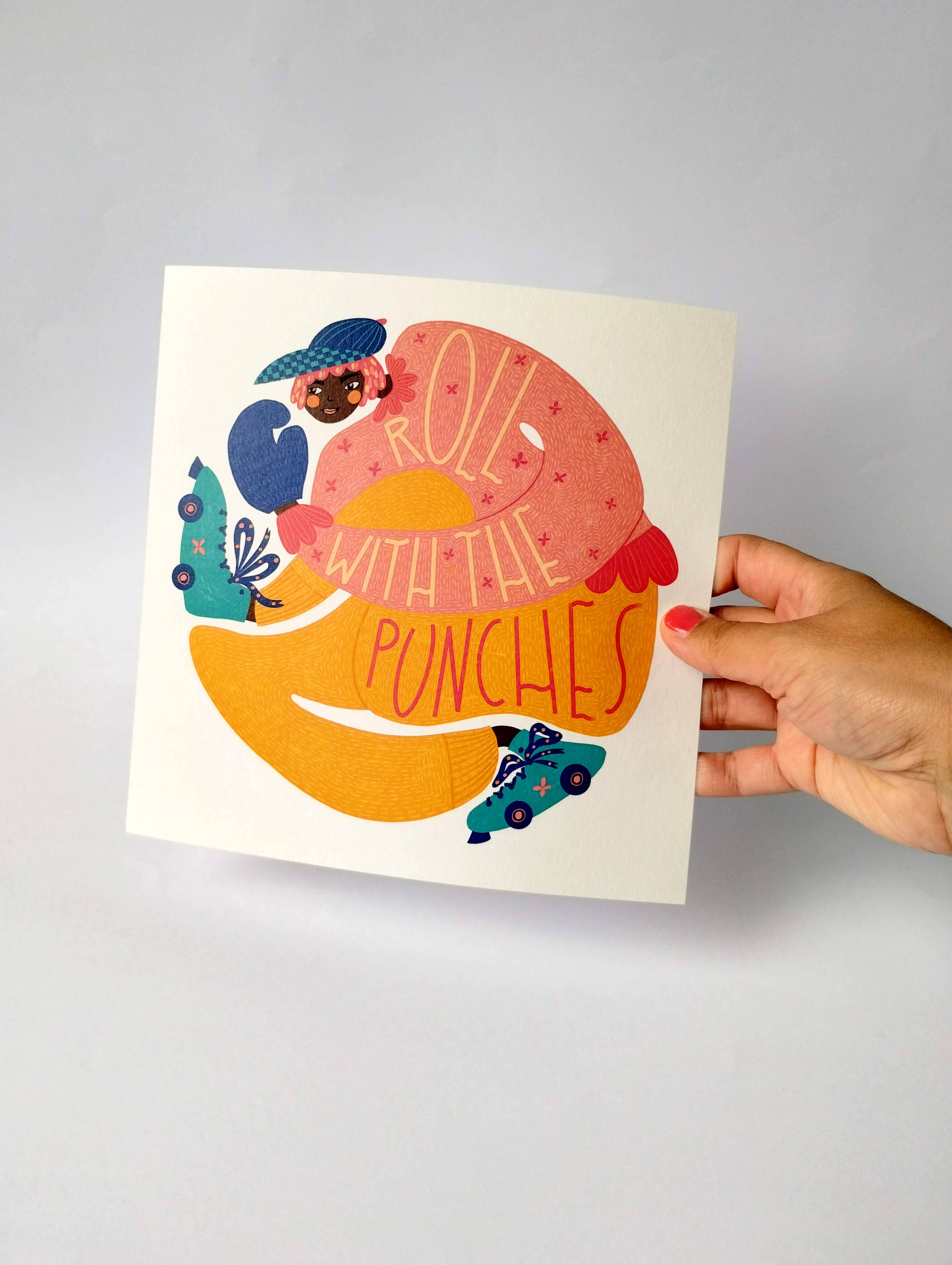 Hand holding a colourful print with a donut illustration and text on a grey background.
