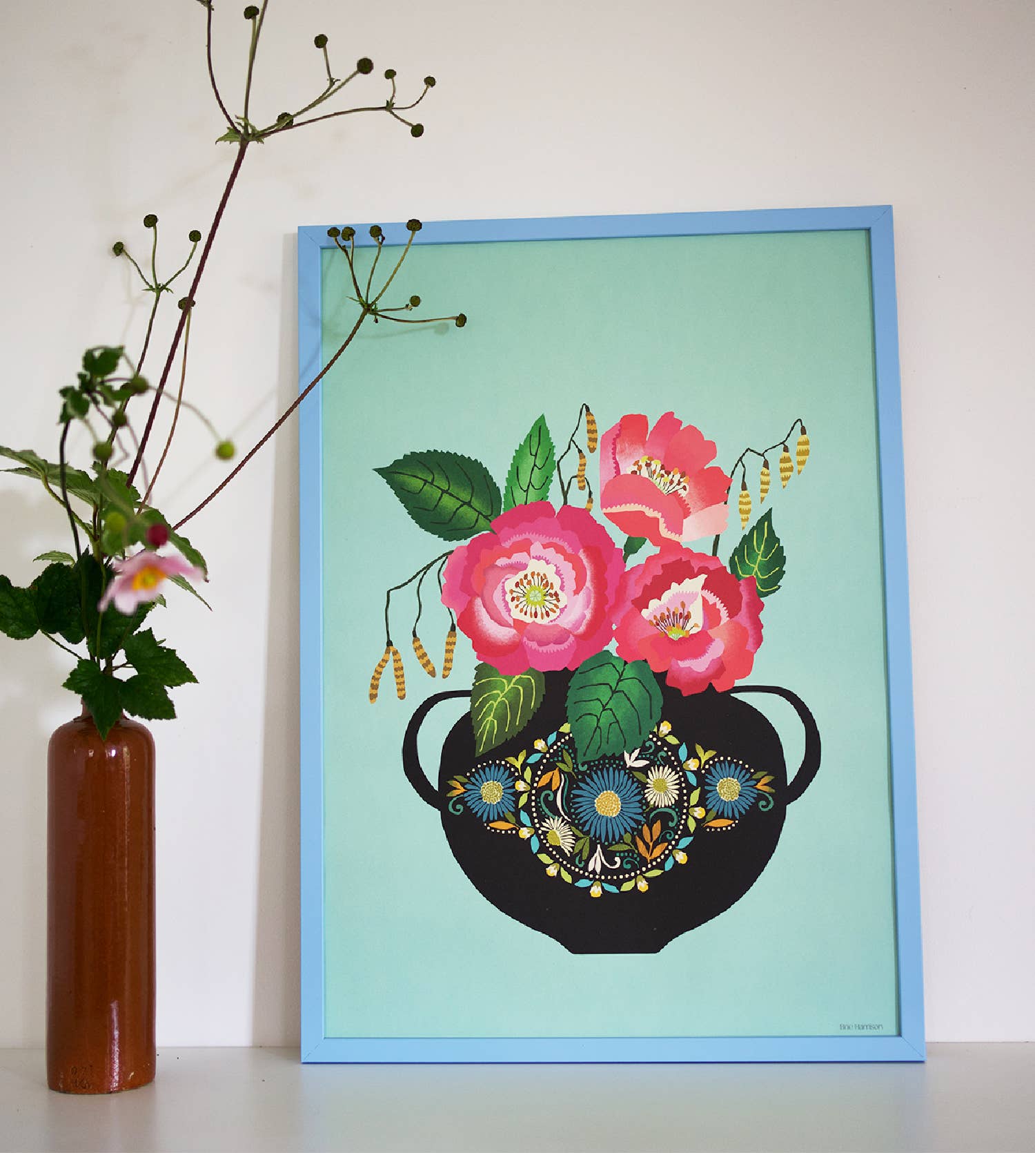 Framed artwork of a teapot with flowers on a light blue background, next to a vase with flowers.