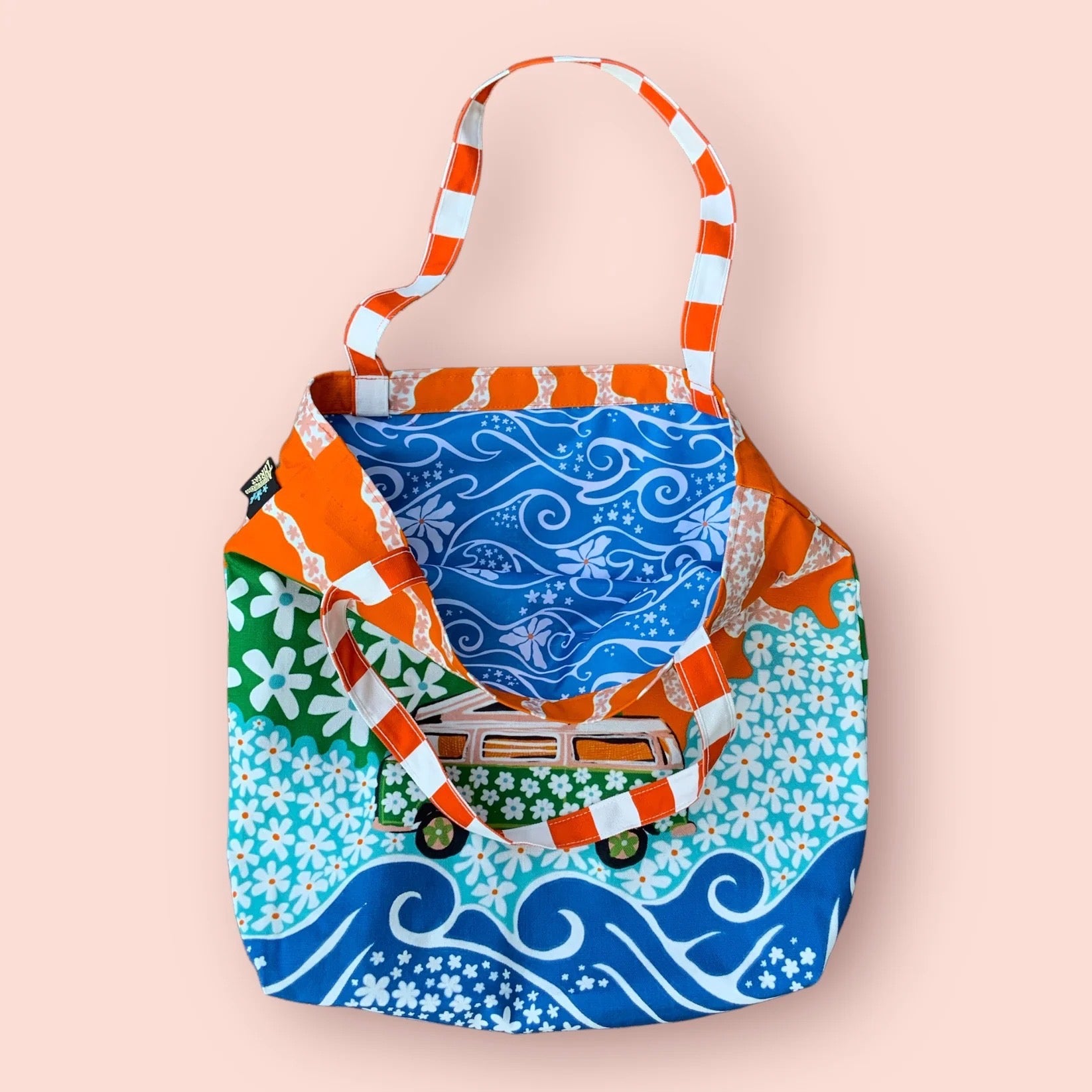 A fabulous, bright and colourful tote bag featuring a pink campervan covered in green daisies on a meadow of blue daisies, a large orange sun and blue waves. This tote bag is made from 100% cotton half panama with retro waterproof lining.